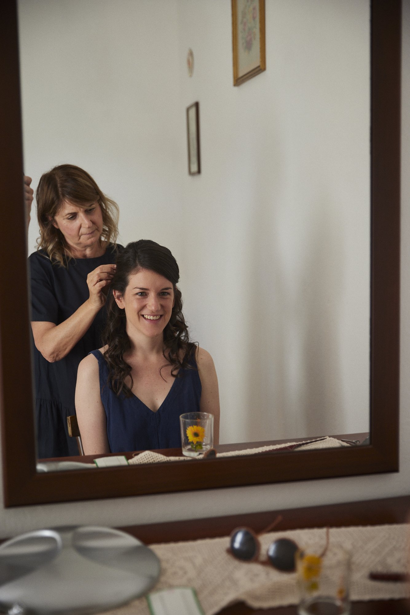 A woman sitting in front of a mirror, smiling as another woman styles her hair.