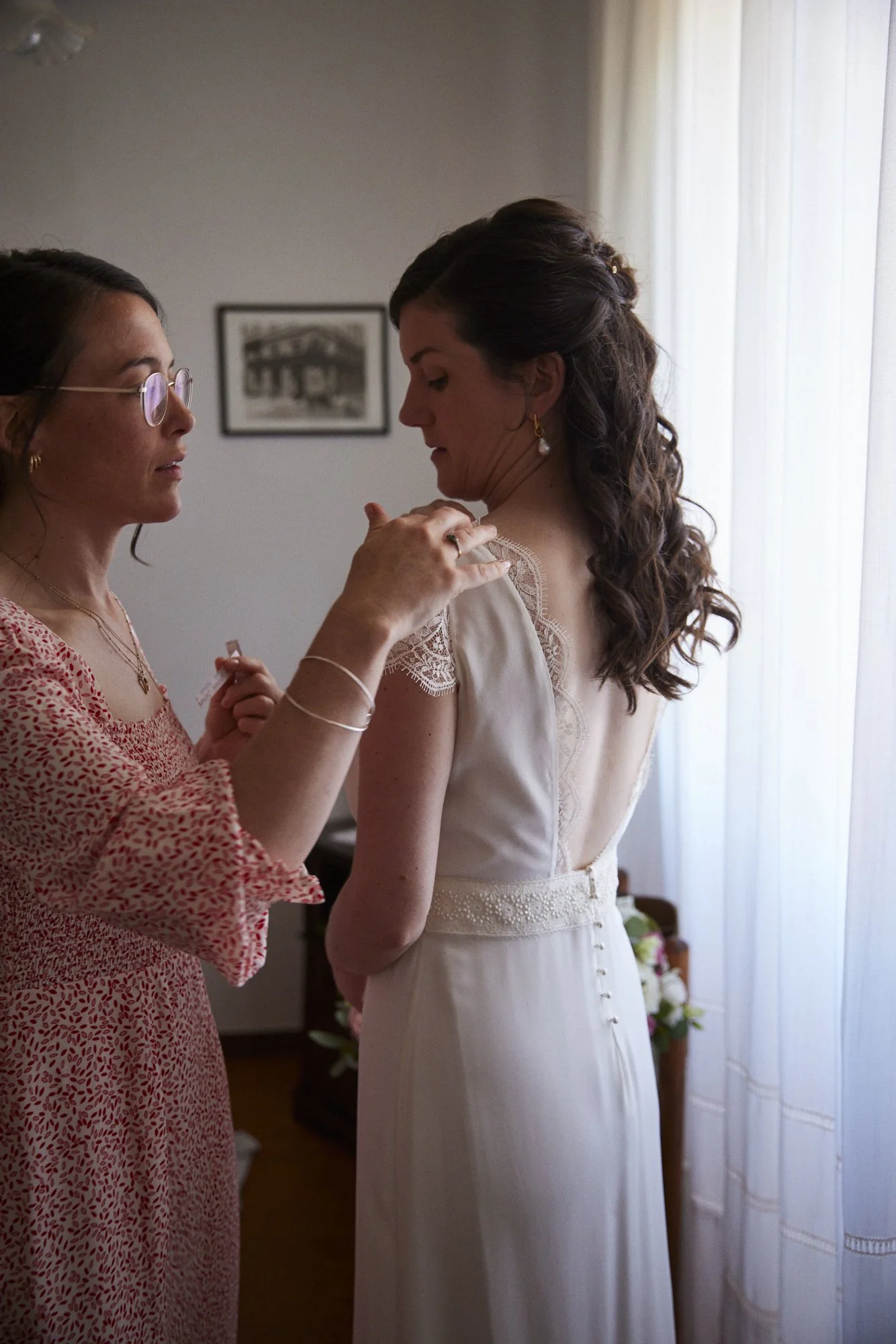 A woman in a wedding dress having her shoulder adjusted by another woman in a patterned dress indoors near a window with curtains.
