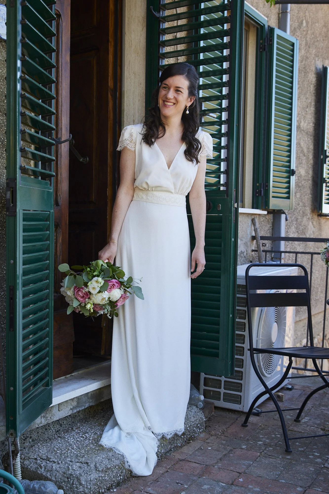A woman in a white dress stands on a porch holding a bouquet of pink and white flowers, smiling.