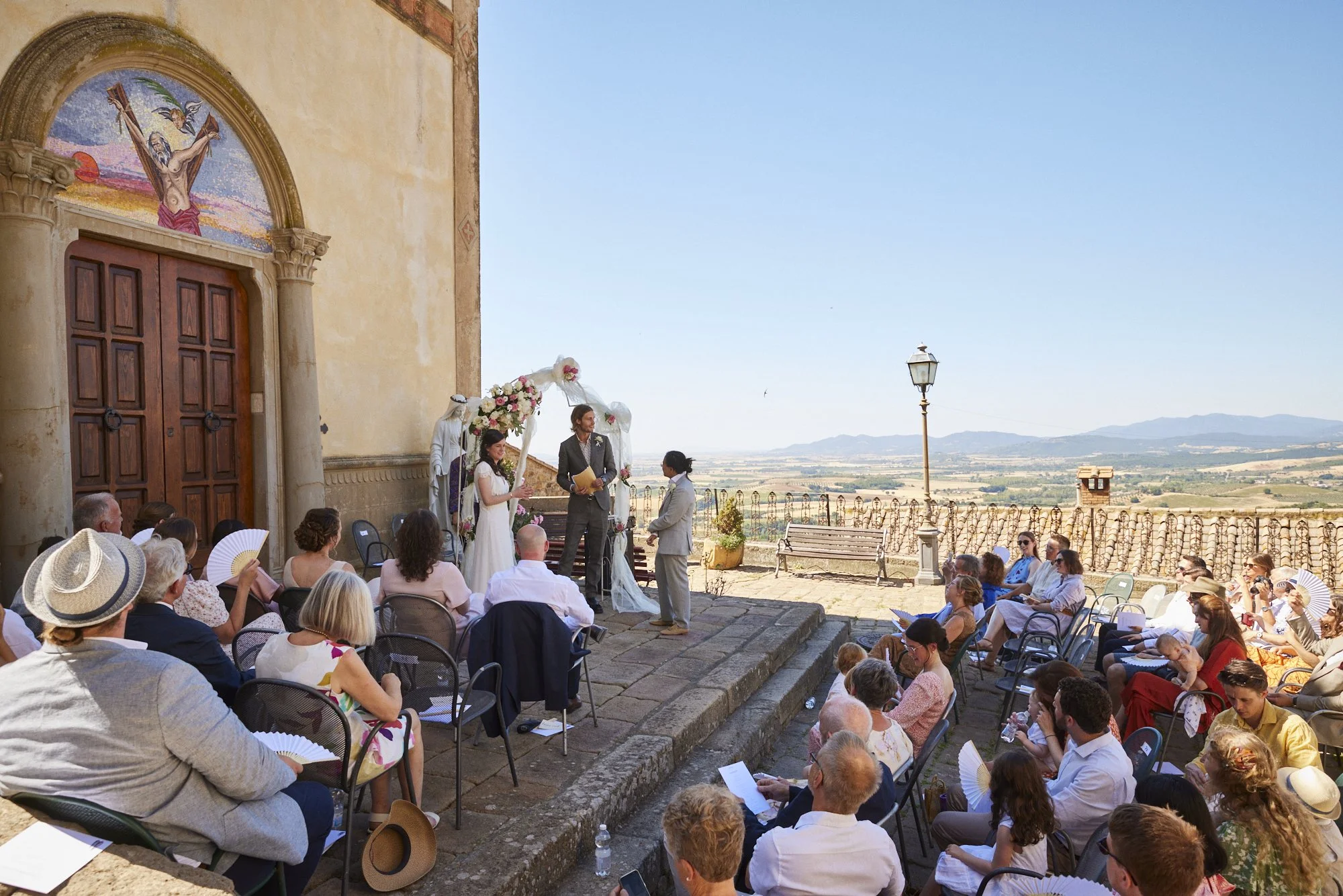 Outdoor wedding ceremony taking place in front of a historic building with a painted archway, with guests seated in chairs on stone steps, and scenic countryside in the background.