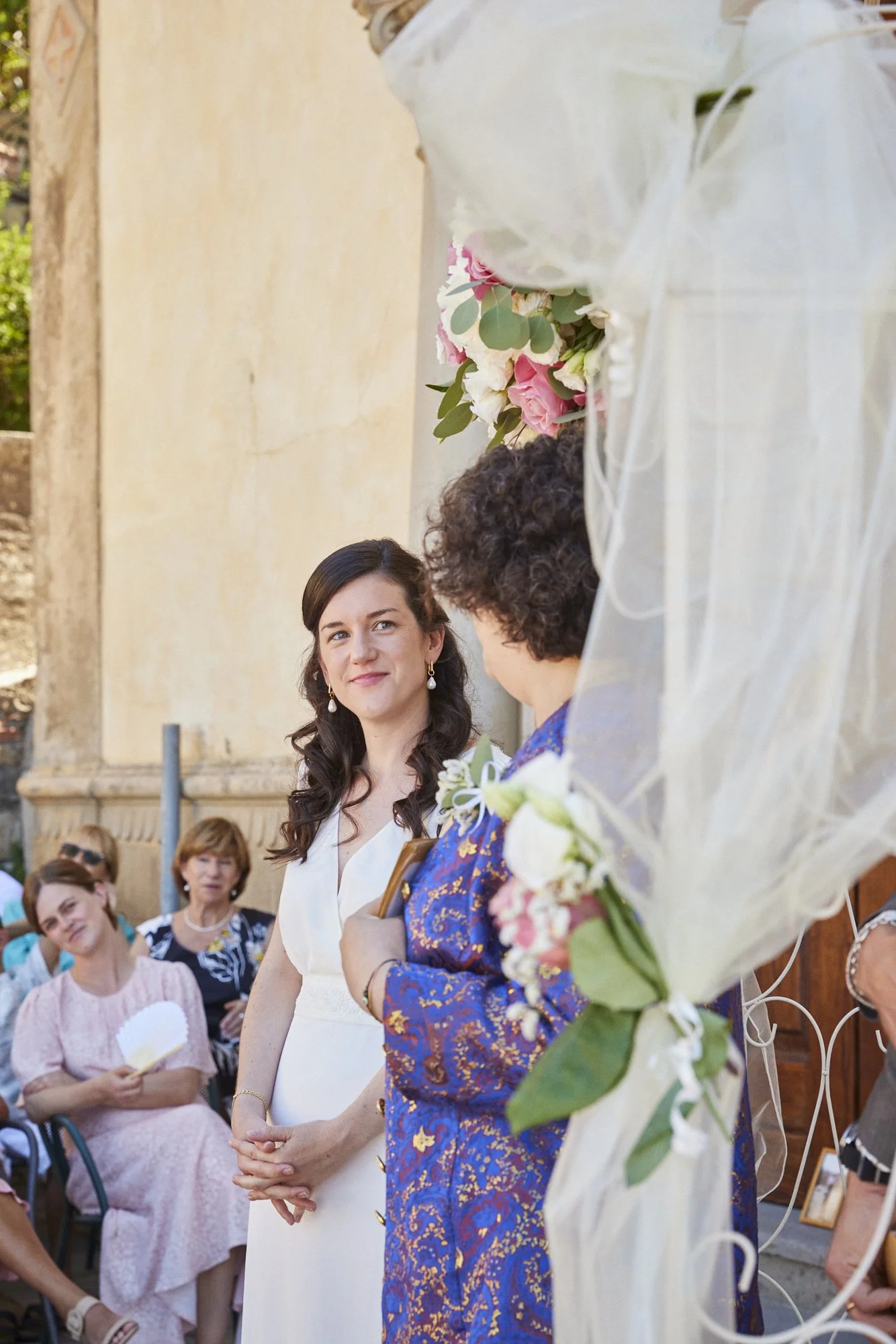 A bride in a white dress looks at an older woman in a blue patterned dress, near an arch decorated with pink and white flowers, during a wedding ceremony outdoors.