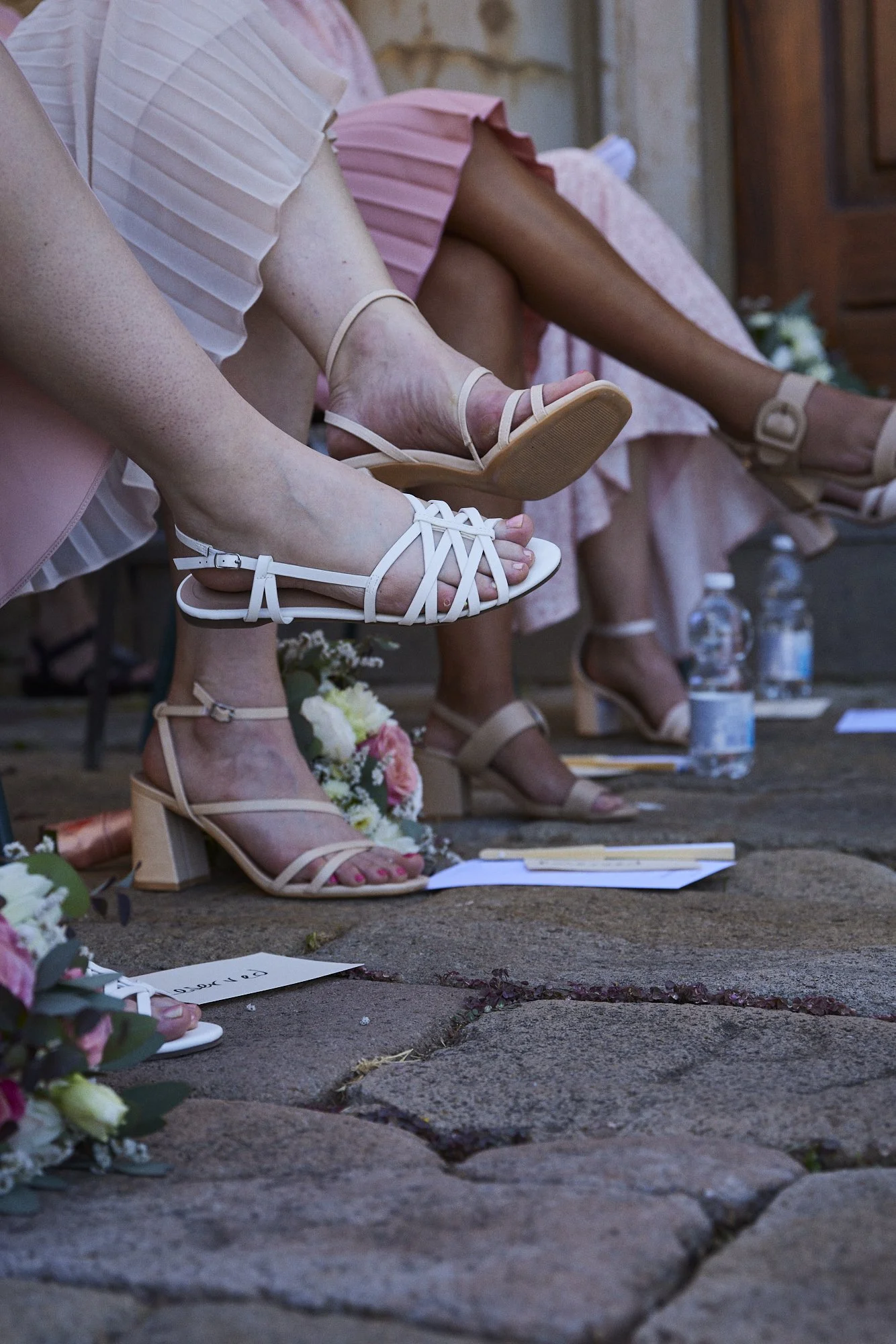 Close-up of women sitting on a bench, wearing pink dresses and beige or white high-heeled sandals, with flowers and water bottles on the ground.