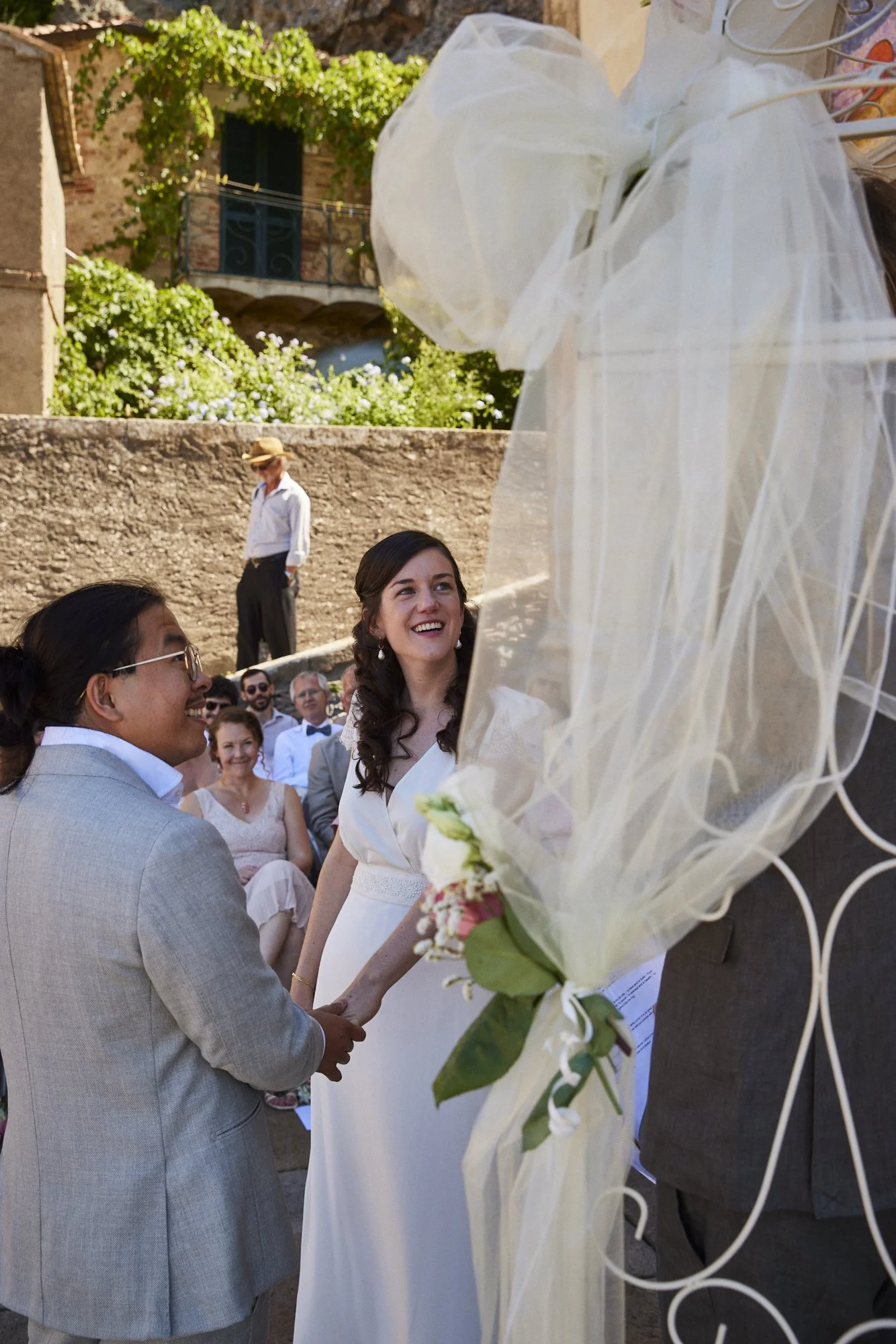 A wedding ceremony outdoors with a bride and groom holding hands, facing each other, surrounded by guests. The bride is smiling, wearing a white dress, and the groom is in a light gray suit. There is a large floral arrangement with sheer white fabric