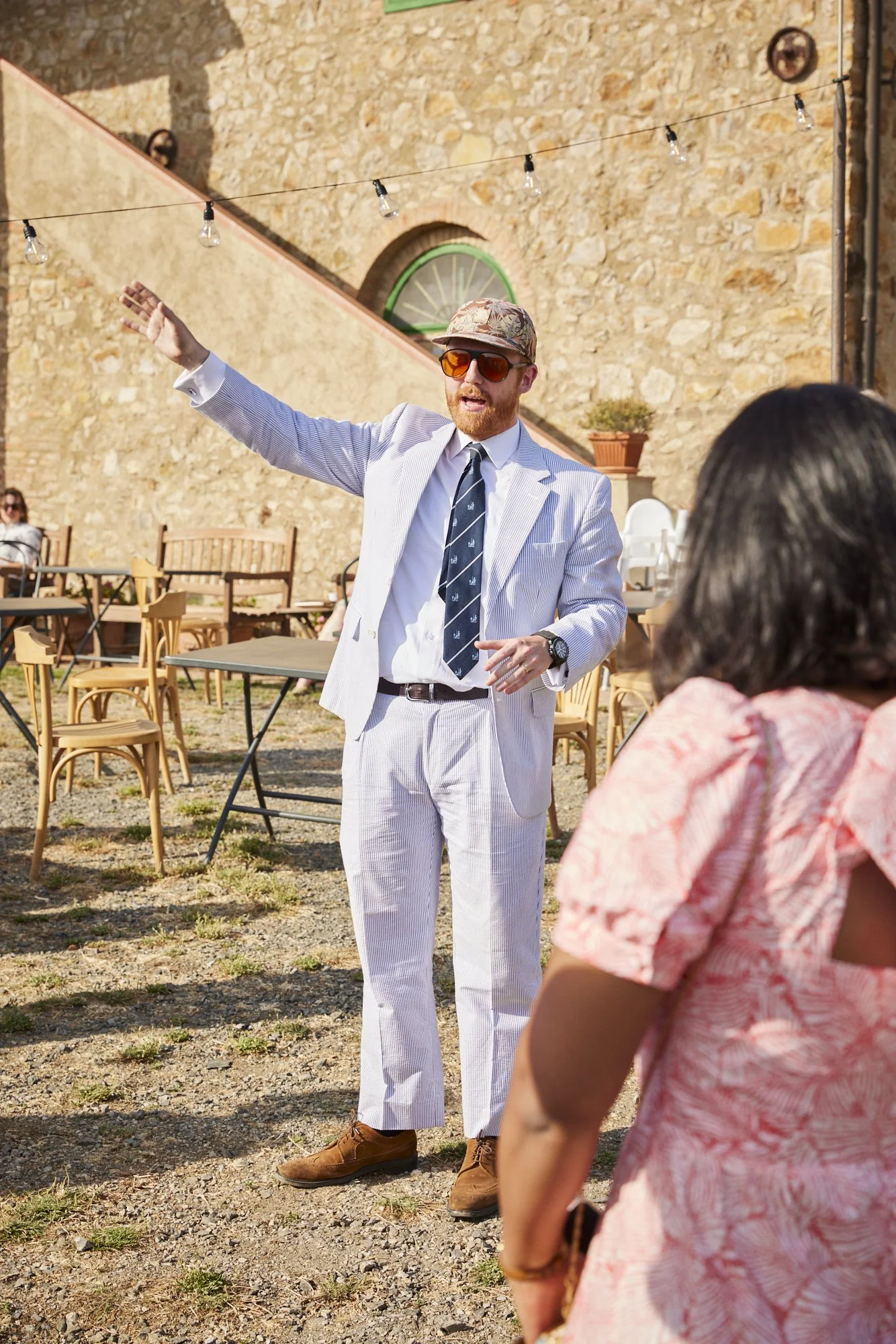 A man wearing a light-colored suit, sunglasses, a hat, and a tie is gesturing with his right arm while speaking outdoors at a gathering. There are chairs and tables behind him, and a stone wall with string lights overhead.