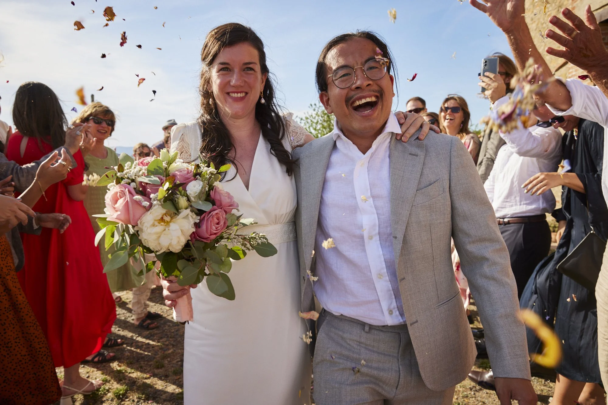 A bride and groom smiling and walking together joyfully, surrounded by friends and family celebrating outdoors on their wedding day.