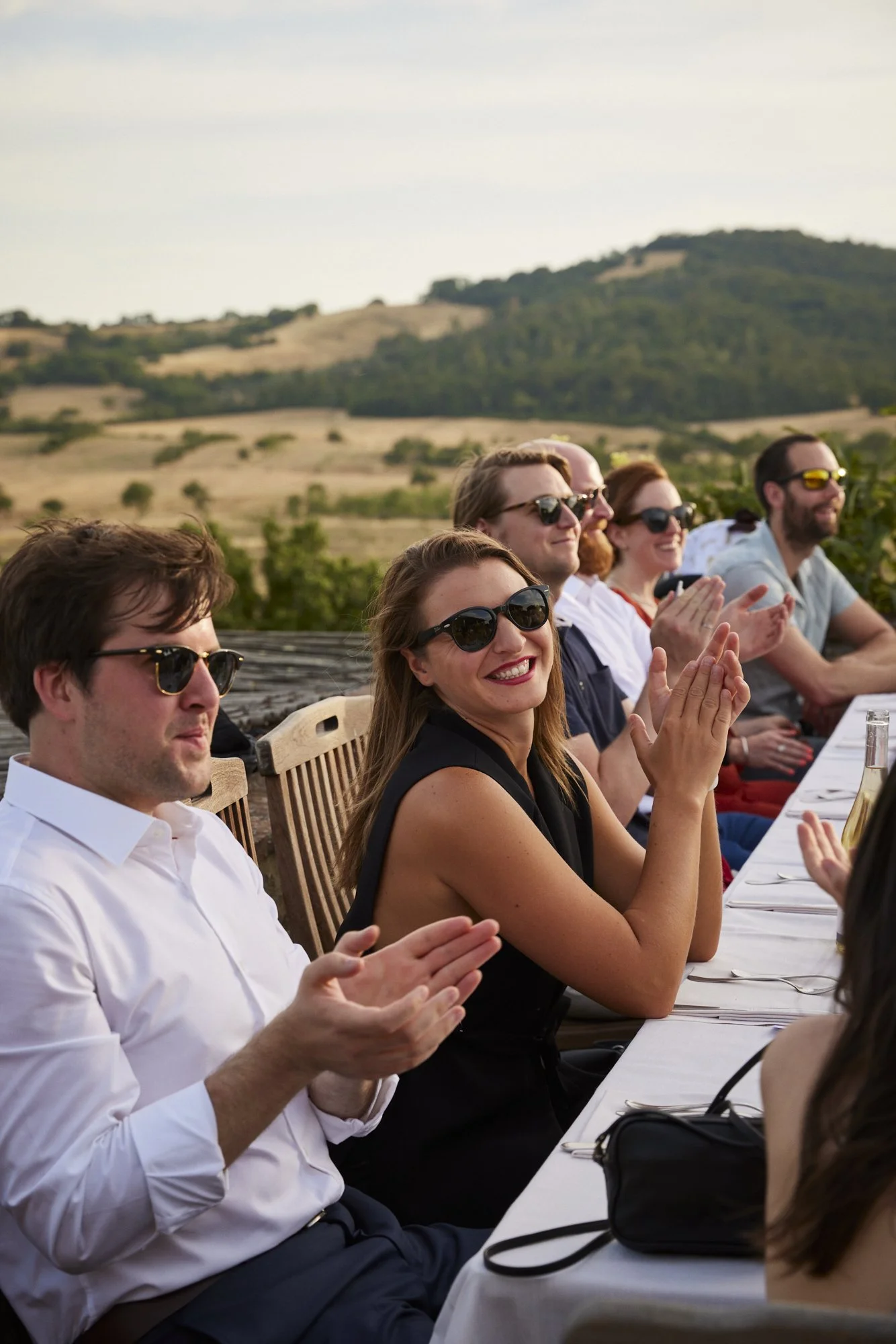 People sitting at a long outdoor table enjoying an event, with a scenic countryside in the background, clapping and smiling, some wearing sunglasses.