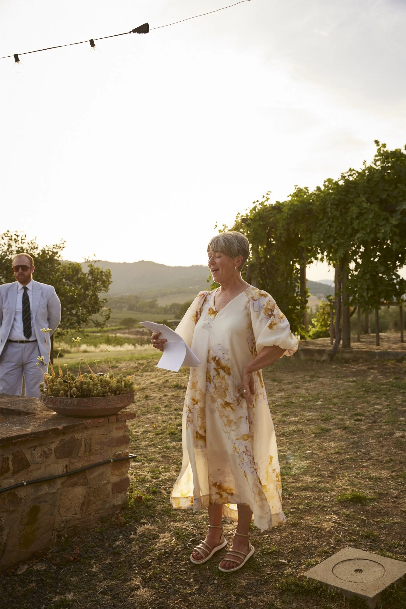 An elderly woman reading aloud while holding papers, dressed in a long beige and gold patterned dress, outdoors during sunset with greenery and a stone structure nearby.