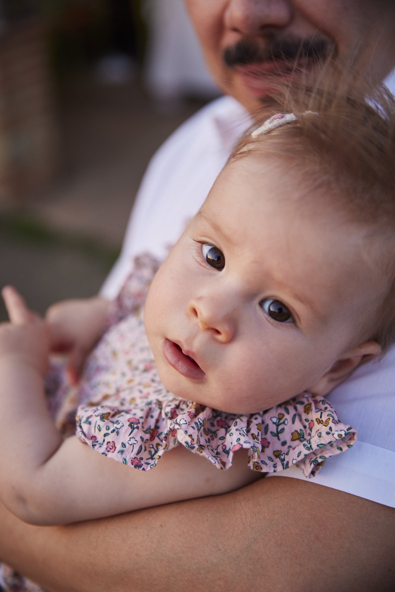Close-up of a baby girl with brown hair and big brown eyes, resting on an adult's arm, with a man smiling behind her, outdoors.