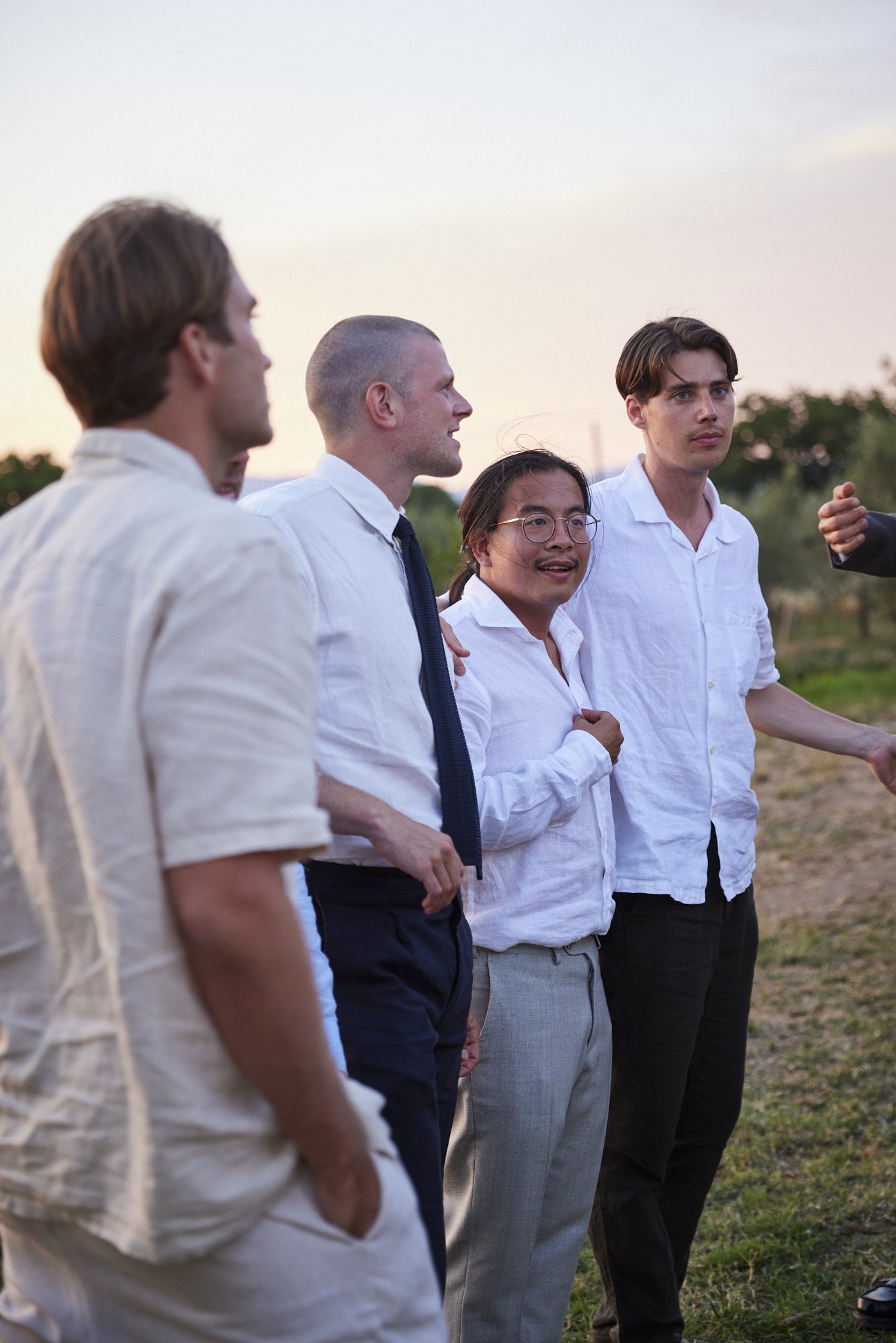A group of five men standing outdoors in a field during sunset, dressed in semi-formal clothing, engaged in conversation.
