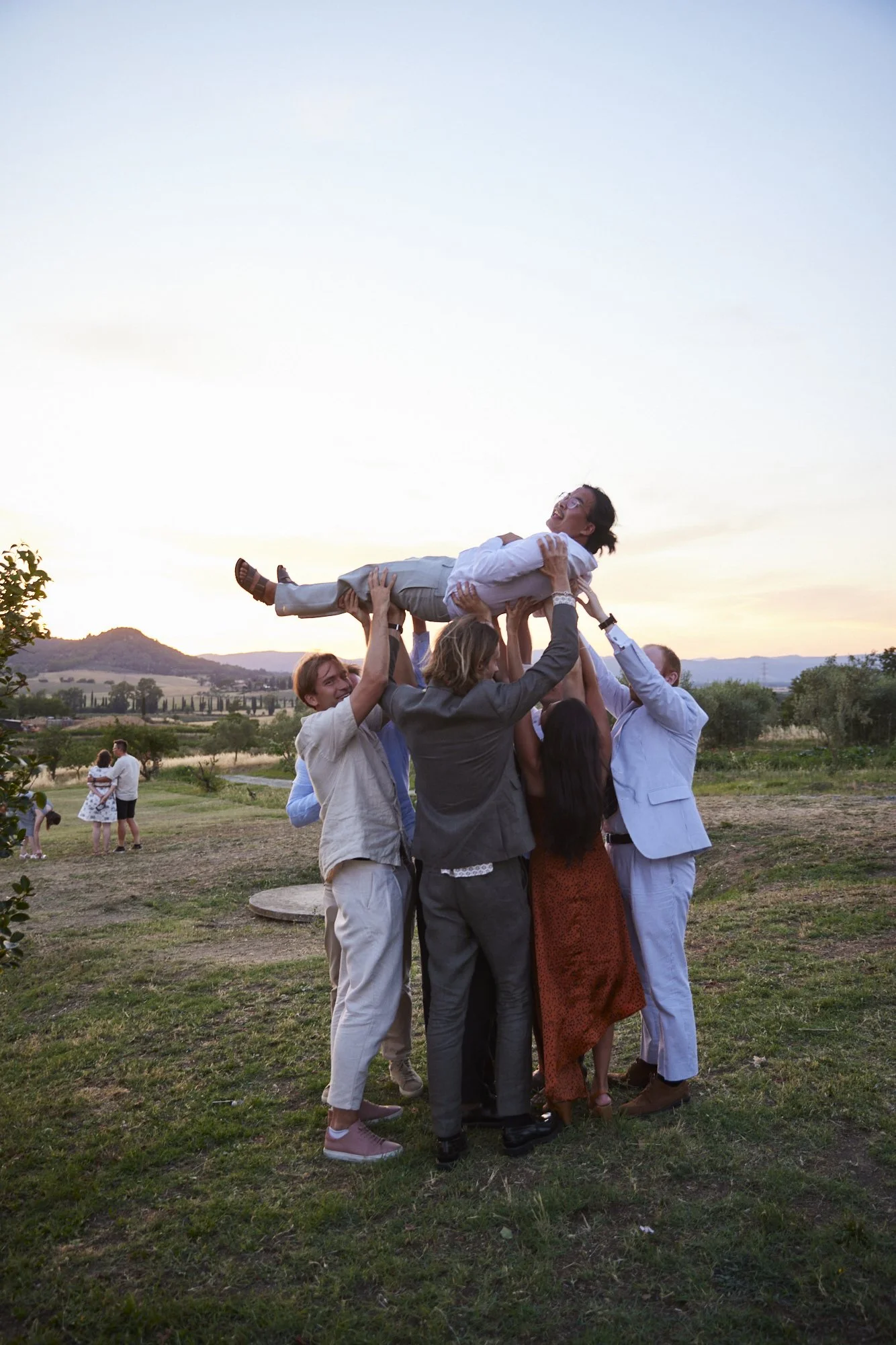 Group of people at an outdoor celebration lifting a woman into the air during sunset.