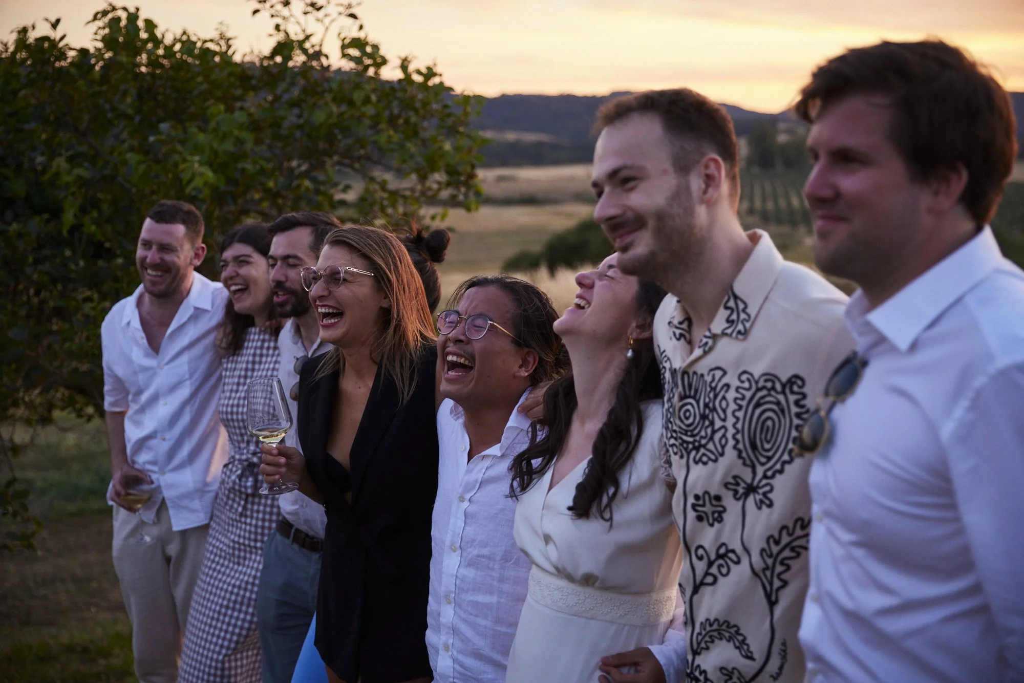 A group of eight people standing outdoors during sunset, smiling and laughing, holding drinks, with fields and trees in the background.