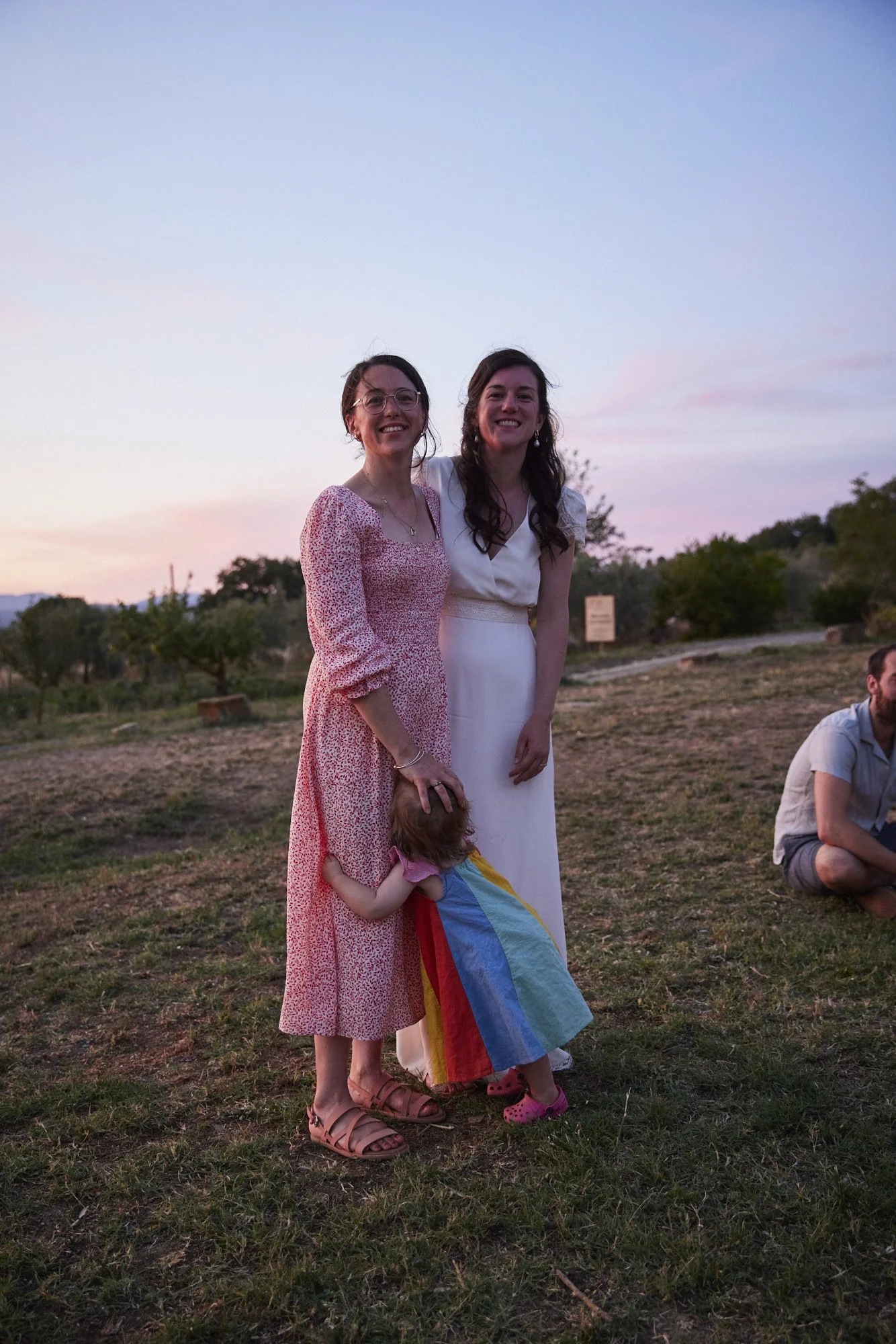 Two women smiling and standing together outdoors at sunset, one wearing a pink dress, the other in white, and a young girl hugging one of the women, wearing rainbow-colored dress and pink shoes.