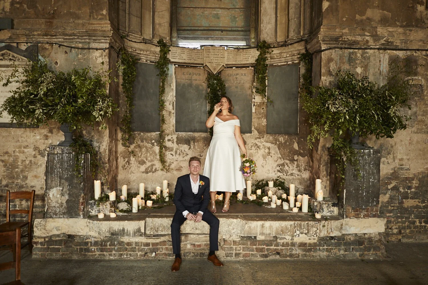 A bride in a white dress and a groom in a dark suit sit together in front of an old, rustic brick wall with candles and greenery decor, with the bride holding a bouquet and standing on a platform.