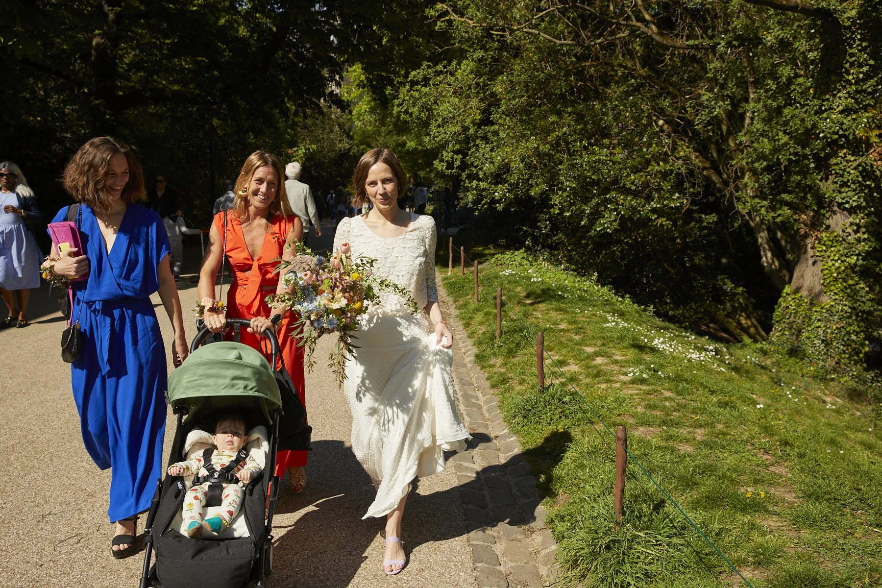 Women in colorful dresses walking outdoors on a sunny day, one holding a bouquet of flowers and a stroller with a young child, in a park with greenery and trees.