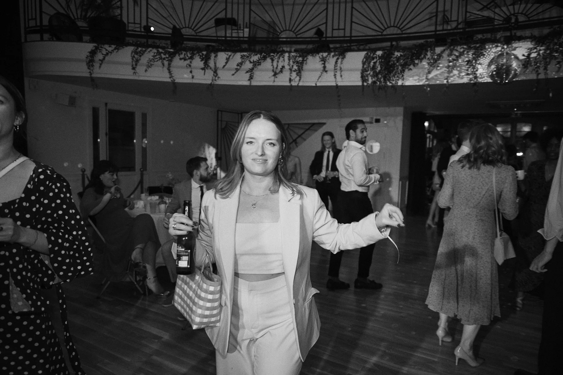 A woman in a light-colored suit holding a drink at a lively social event, with other guests mingling in the background.