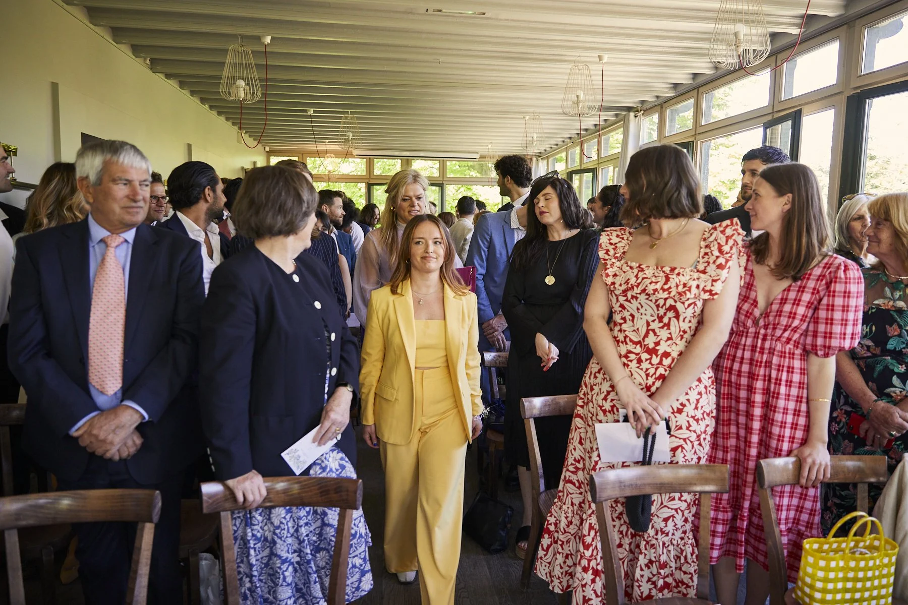 Group of diverse people standing and conversing in a bright, indoor space with large windows and modern decor, during a social or professional gathering.
