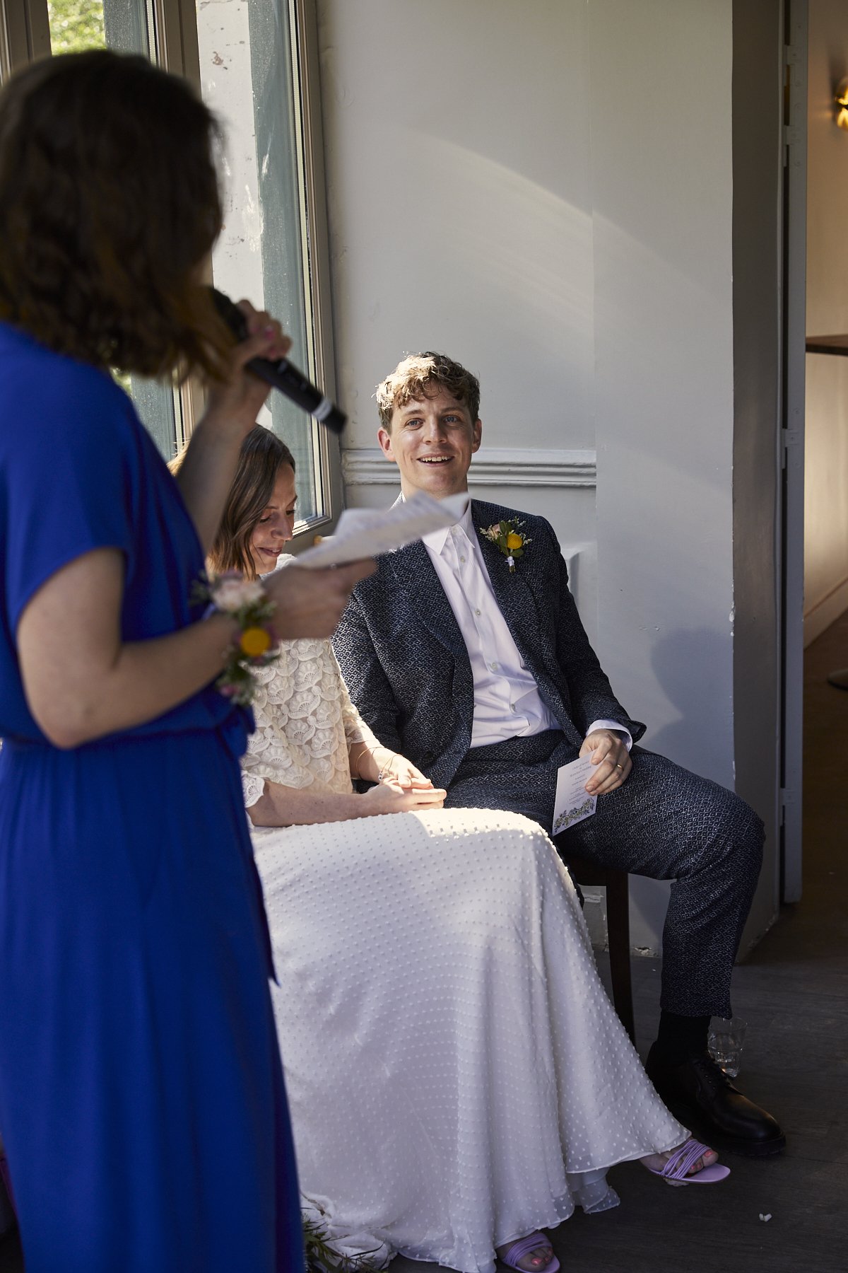 A woman in a blue dress is giving a speech during a wedding ceremony, with the bride and groom sitting side by side and listening, standing near a window with sunlight