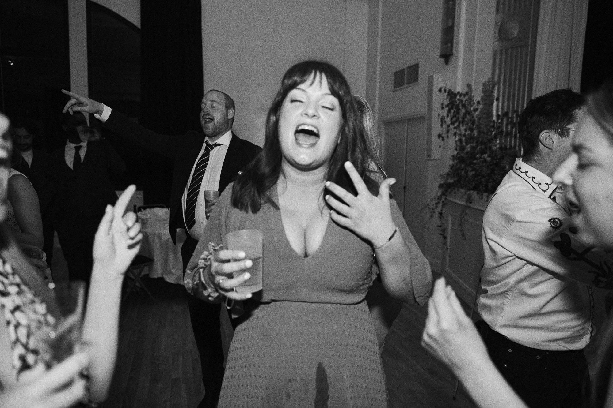People dancing and socializing at a party or celebration indoors. A woman in the center is laughing with a drink in her hand, surrounded by other party-goers.