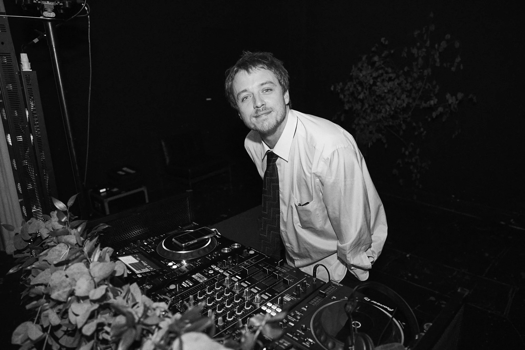 A man wearing a white dress shirt and dark tie standing behind DJ equipment with turntables and a mixer, in a dark setting with plants visible in the background.