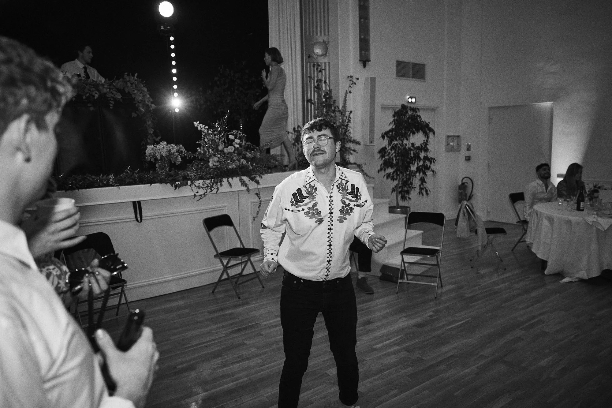 A man dancing at a social event in a banquet hall, with other guests seated at tables and a woman singing into a microphone on stage in the background.