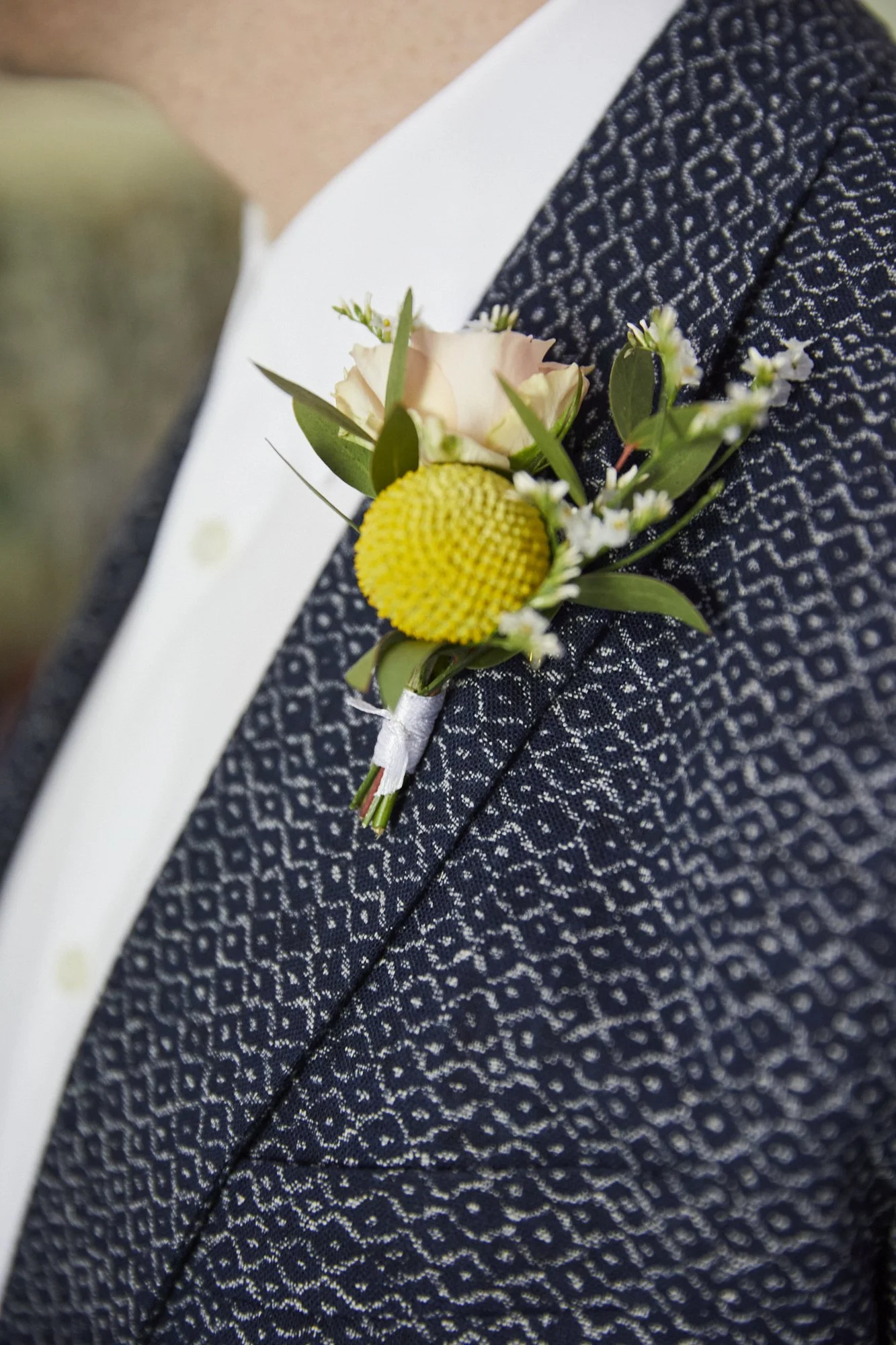 A close-up of a man's suit jacket with a pocket square, paired with a boutonniere consisting of a light pink rose, yellow billy button flower, small white flowers, and green foliage.