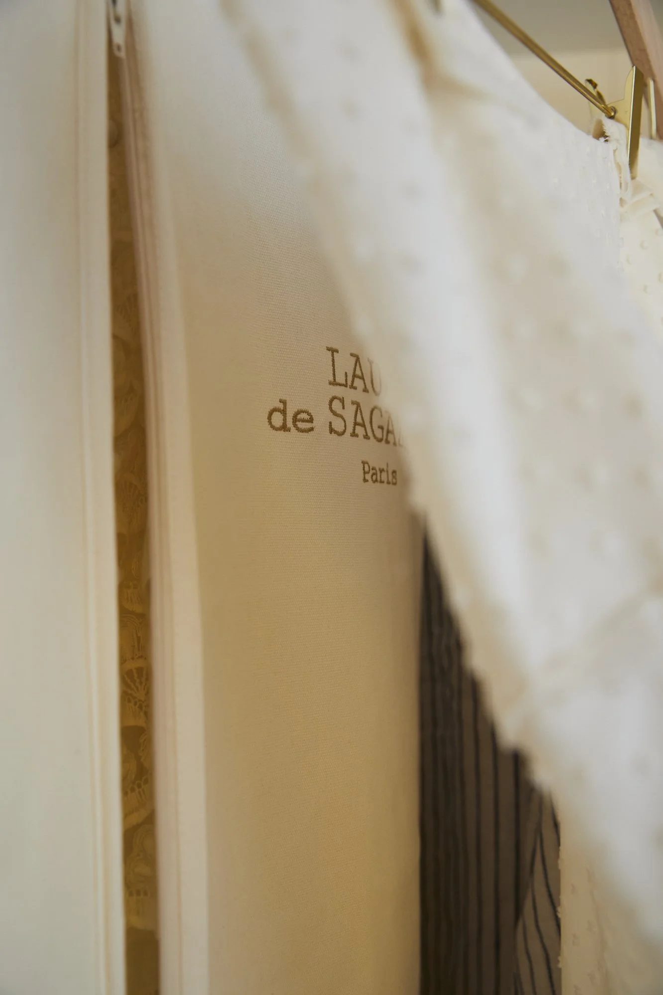 A close-up of a white fabric with a gold-colored clip holding it, revealing a beige-colored framed artwork or menu with "LAD de SAGAL Paris" written on it. The background includes a glimpse of patterned fabric.