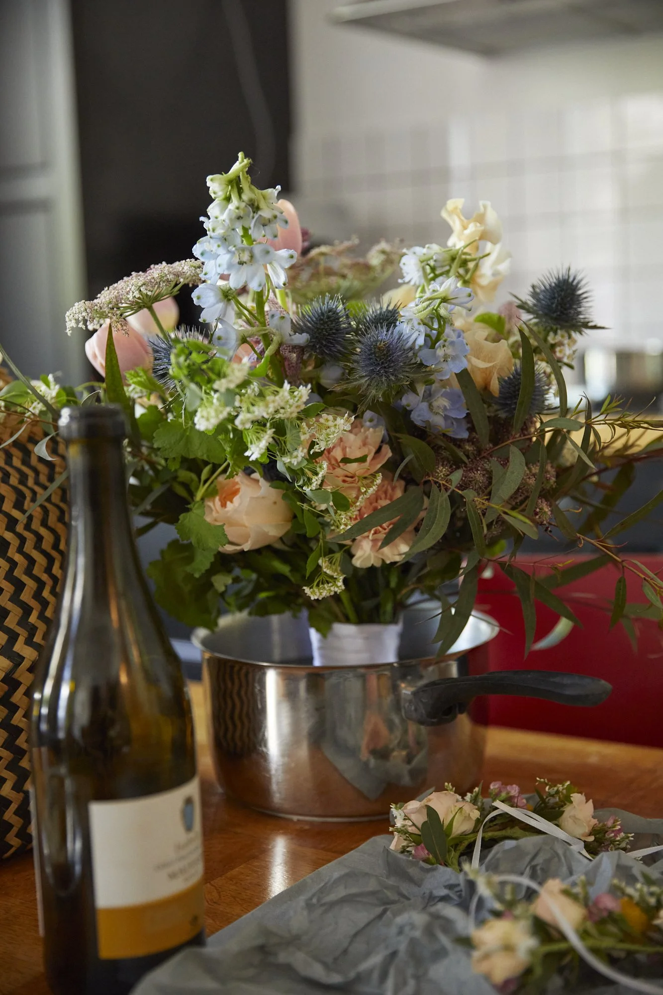 A bouquet of mixed flowers in a silver pot on a wooden table, with a wine bottle and a small floral arrangement in wrapping paper nearby.