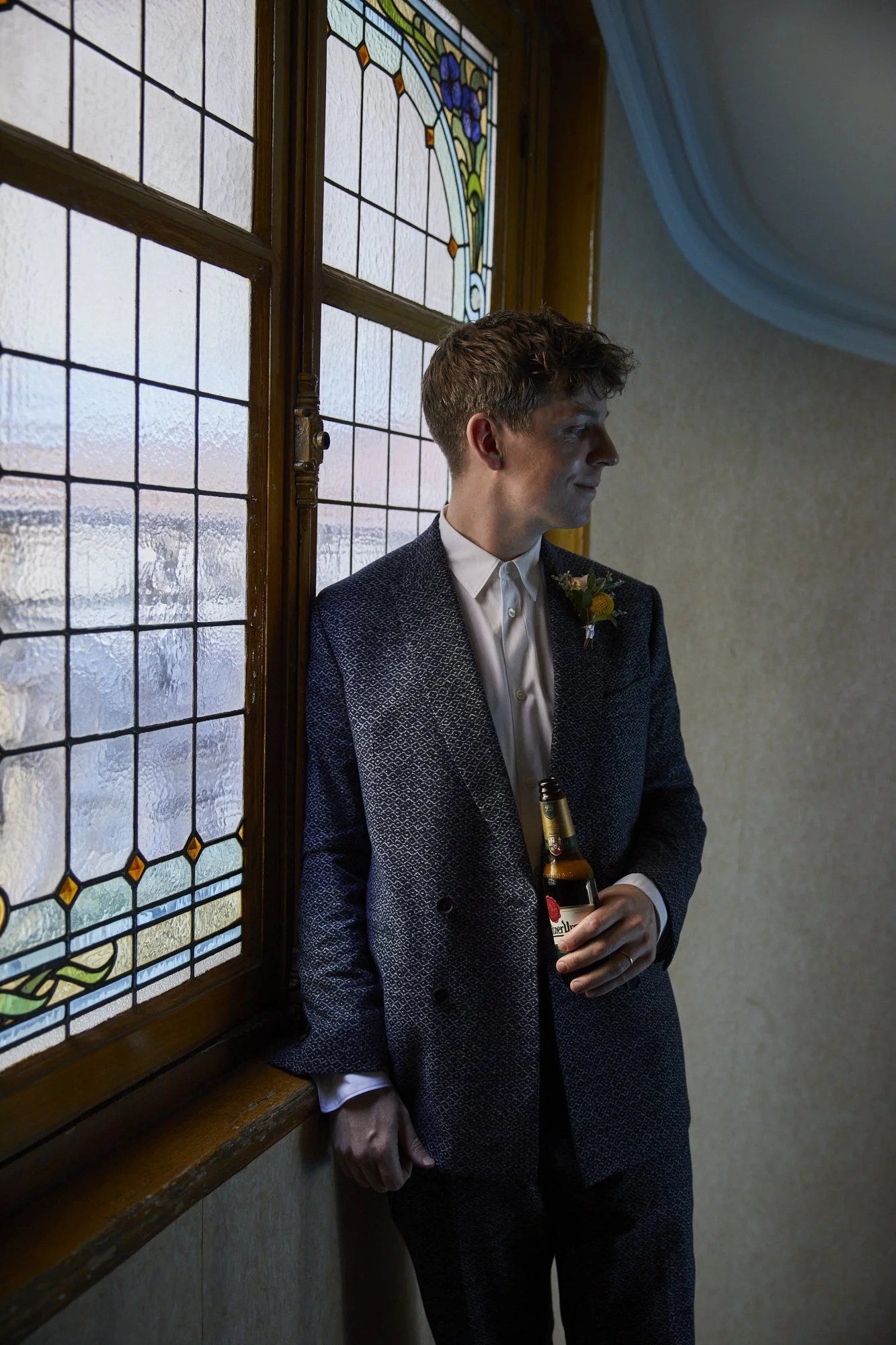 A young man in a suit holding a beer, standing by a window with stained glass and textured glass panels.