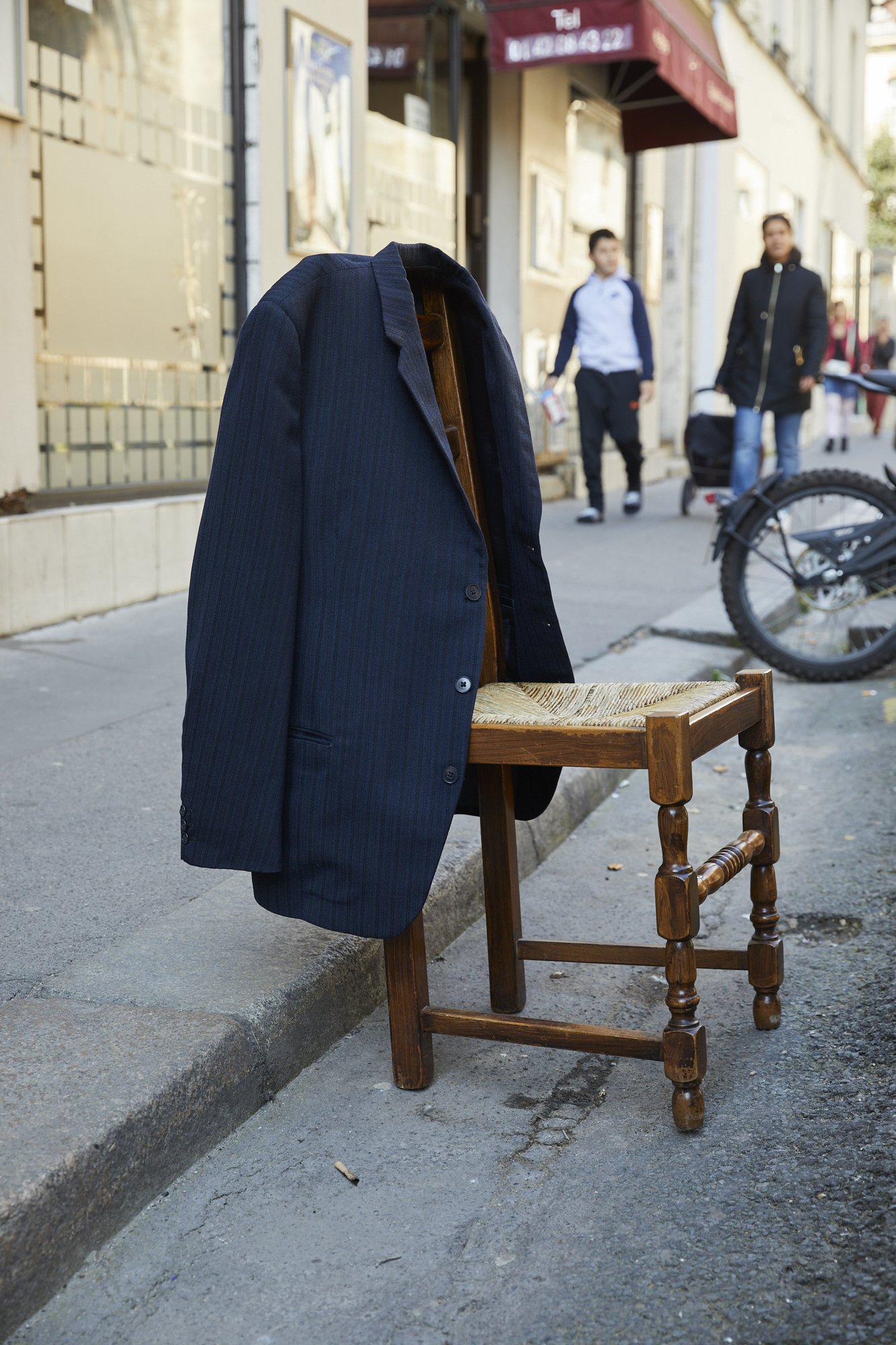 A navy blue blazer draped over a wooden chair placed on a sidewalk in an urban street scene, with pedestrians walking in the background.