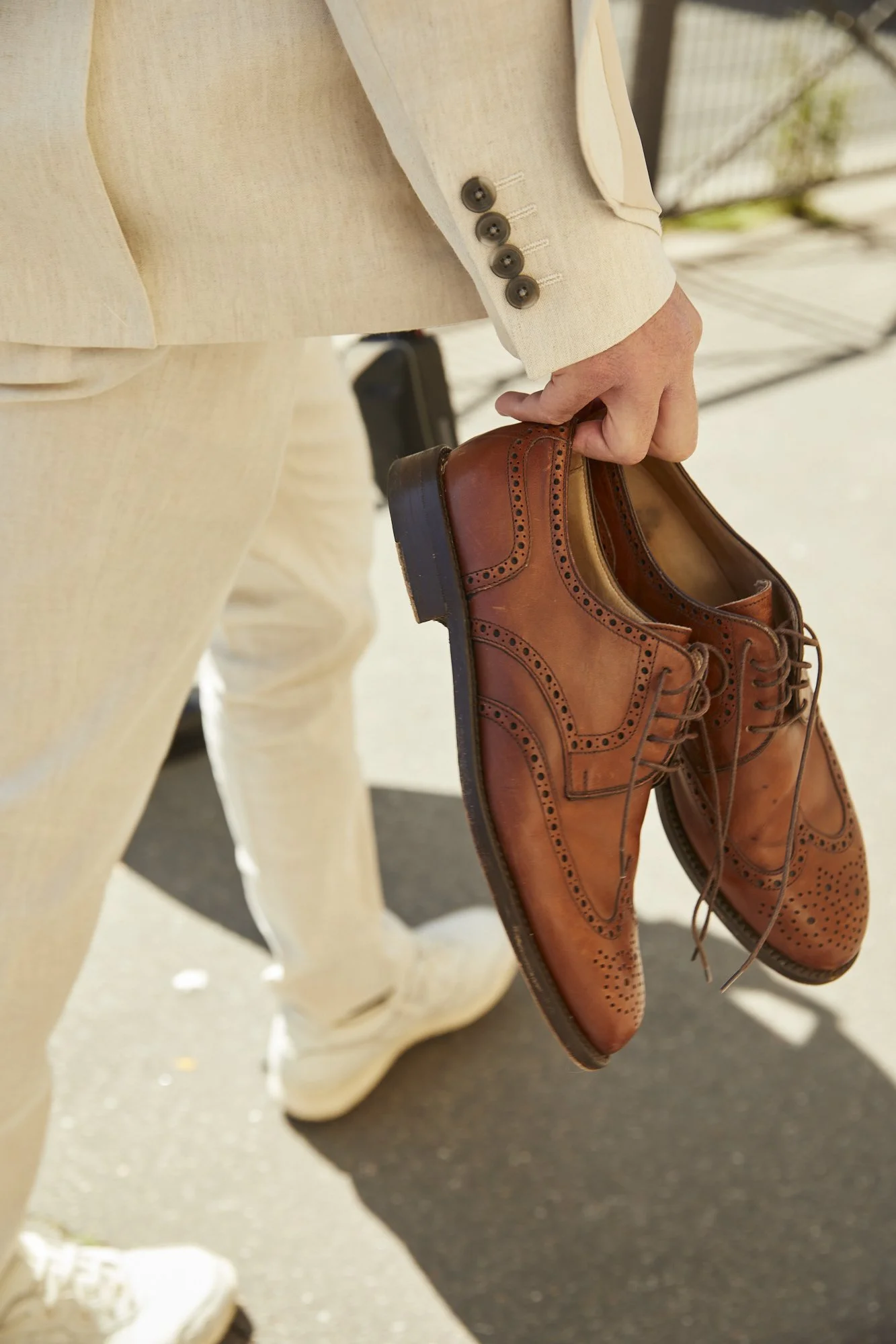 Person dressed in beige suit holding a pair of brown leather dress shoes.