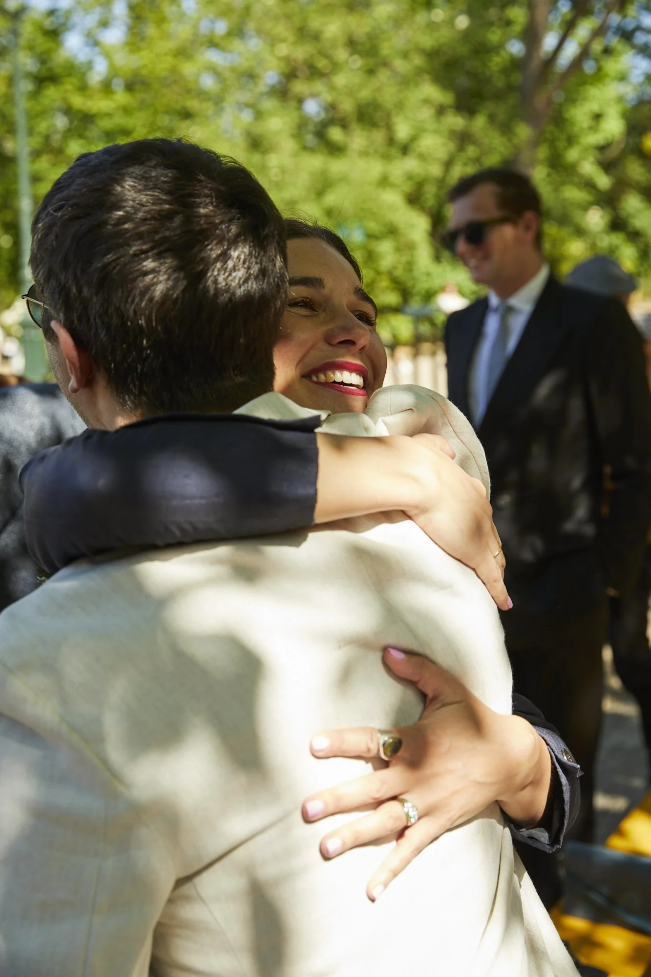 A woman with a big smile hugging a man outdoors, with other people in the background, surrounded by green trees.