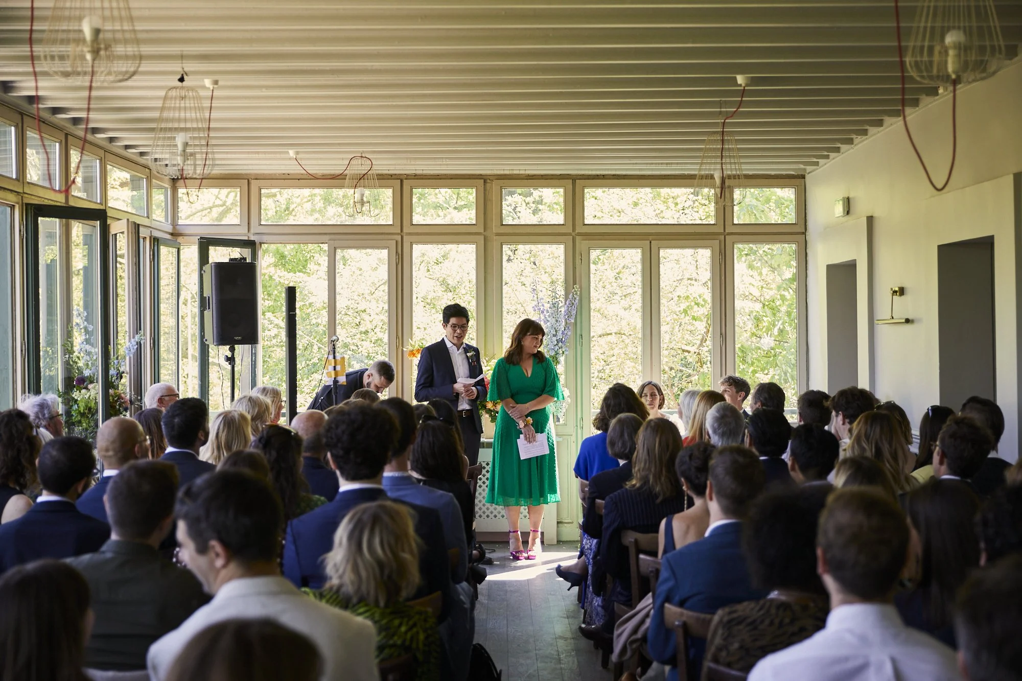Wedding ceremony in a bright, glass-walled room with many guests seated and two people standing at the front, likely the officiant and a witness, during daytime with sunlight streaming through the windows.