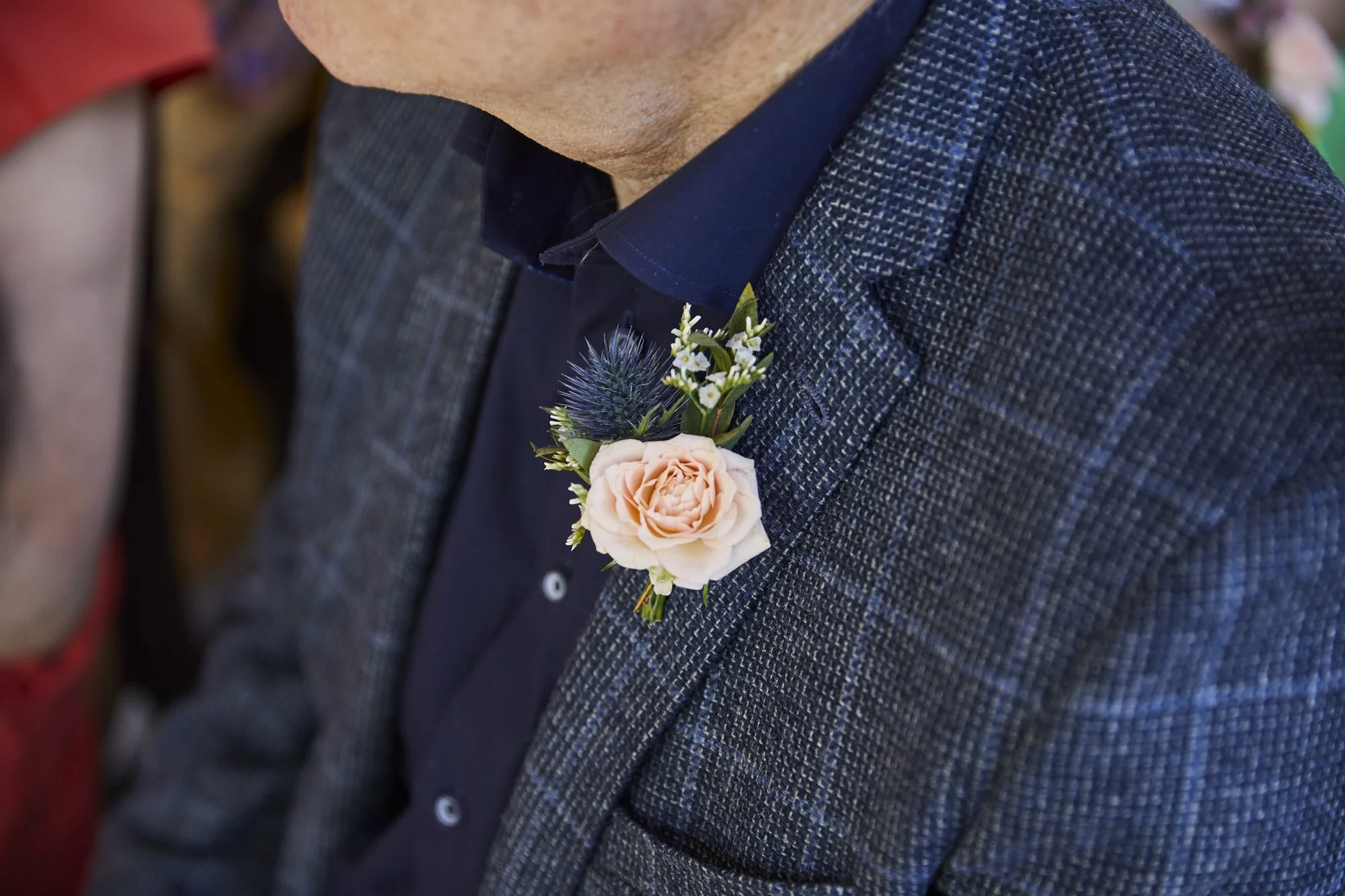 A close-up of a man wearing a dark blue shirt and a gray checkered blazer with a boutonnière featuring a pale pink rose, blue thistle, and small white filler flowers on his lapel.