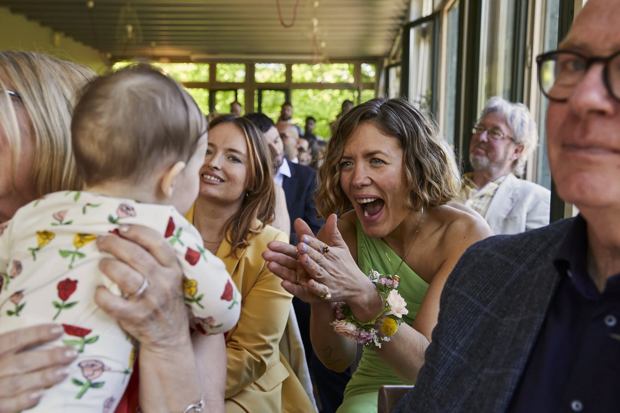 A woman in a green dress enthusiastically talking to a child during a gathering, with other adults seated nearby, some smiling and observing in a sunny room with large windows.