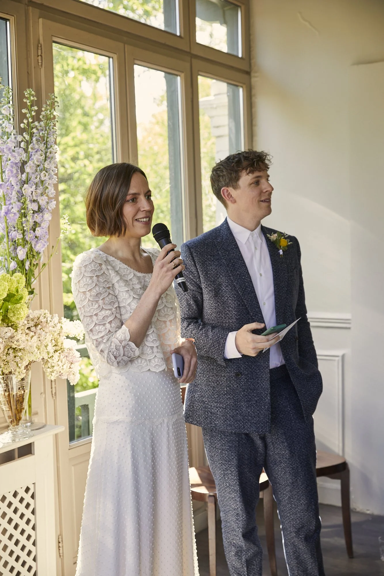 Woman in a white lace dress speaking into a microphone while standing next to a man in a suit at a wedding or formal event near large windows with flowers in the background.