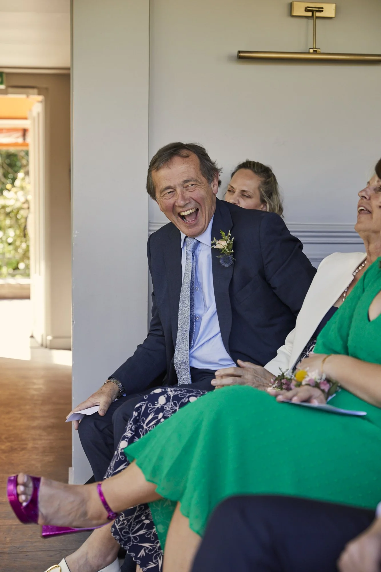 A man in a dark suit laughing with other people seated beside him at a social gathering or ceremony.