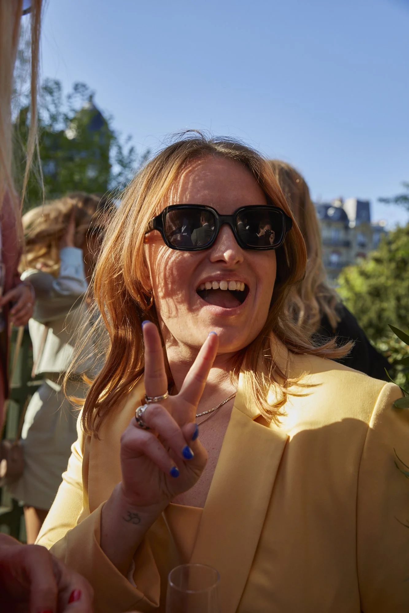 A woman with red hair wearing black sunglasses making a peace sign and smiling at an outdoor gathering.