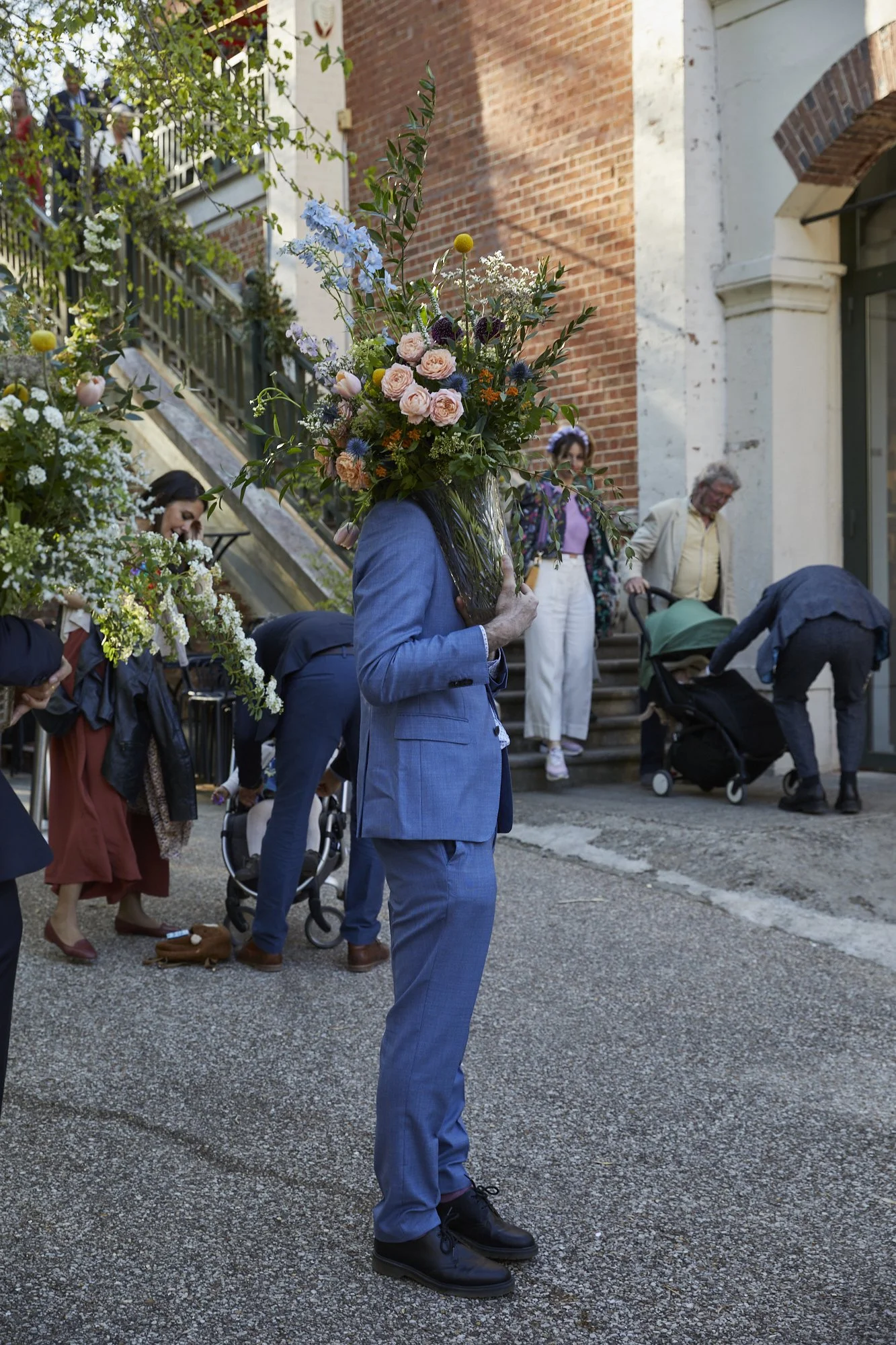 A person in a blue suit with a large bouquet of flowers as a head, standing outdoors on a city street near a brick building, with several people in the background.