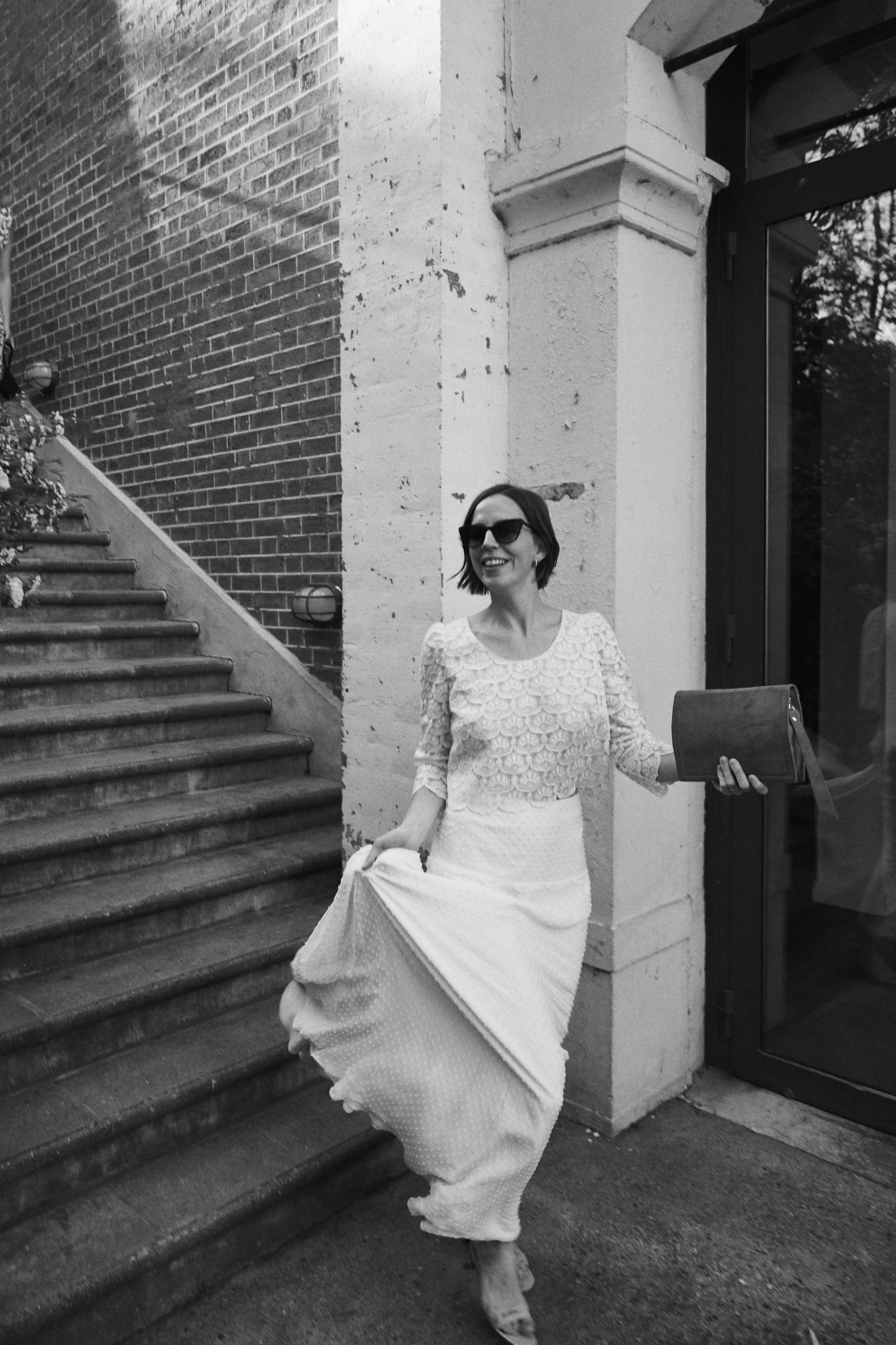 A smiling woman wearing sunglasses and a lace top, holding a clutch and lifting her long skirt while walking down stairs outside a building.