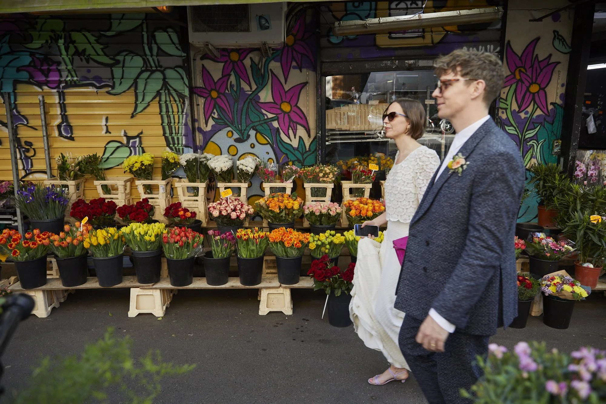 A man and a woman, dressed formally, walking past a flower stand with colorful flowers and graffiti artwork in the background.