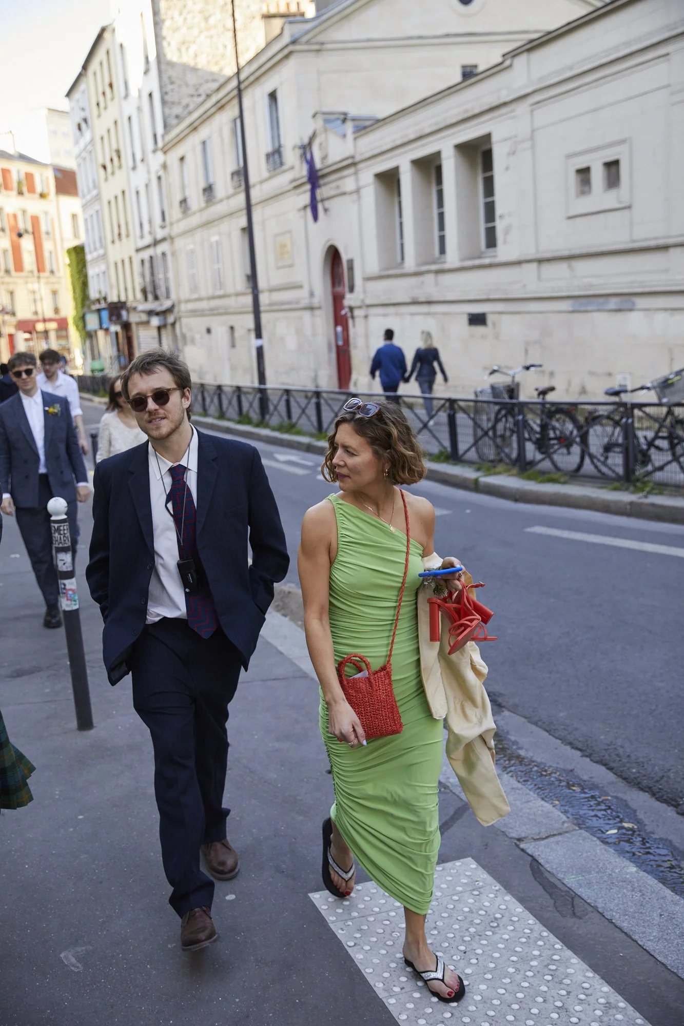 Woman in a green dress walking on a city street with a man in a dark suit, several other people in the background, and buildings lining the street.