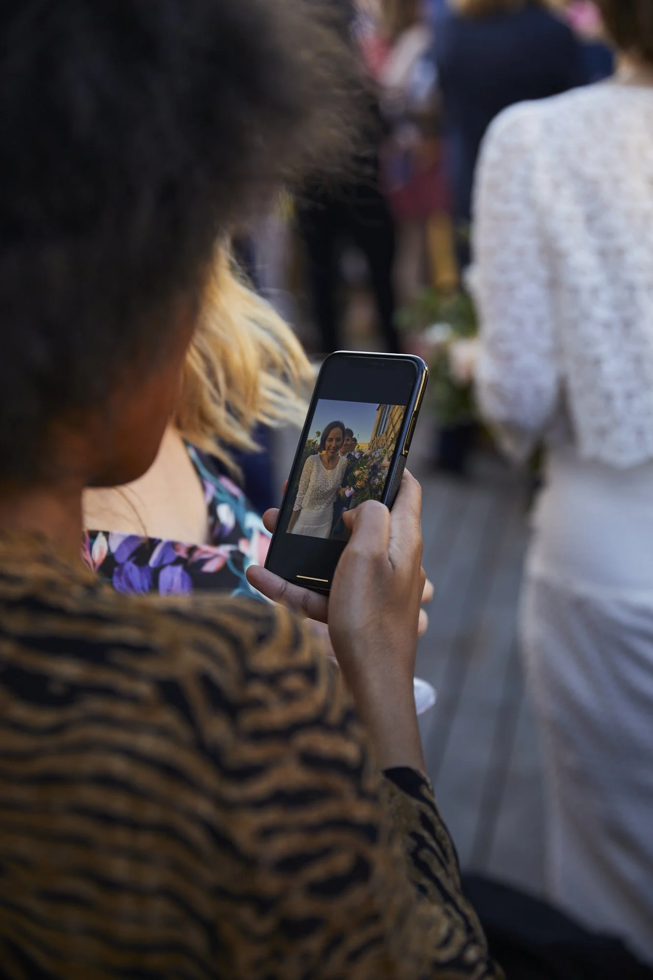 Person taking a photo of a woman holding a bouquet at an outdoor event.