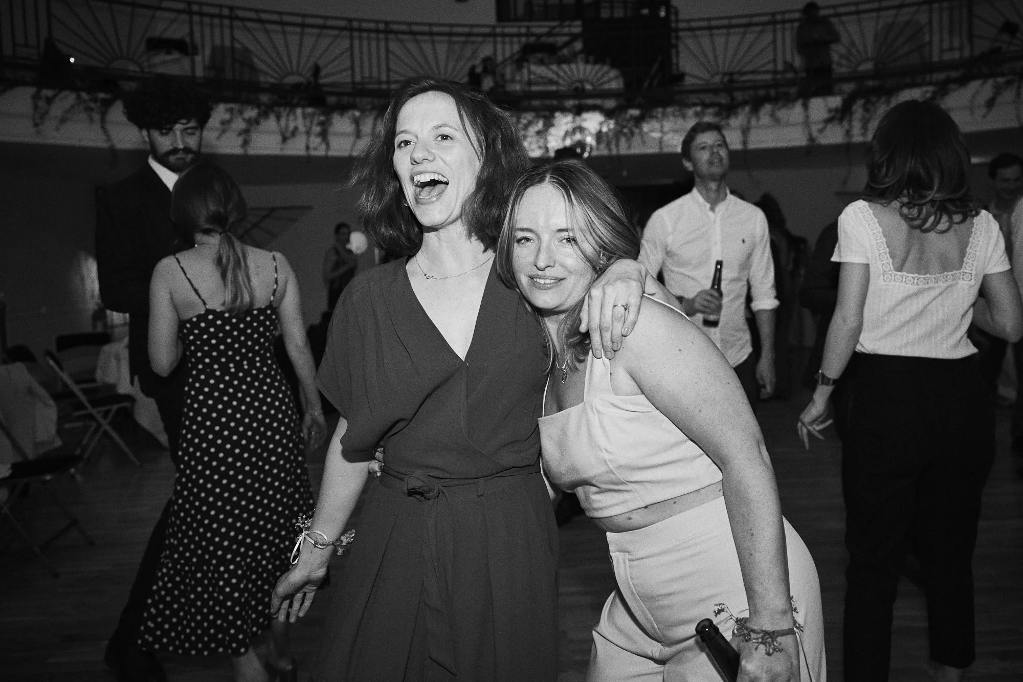 Two women dancing and smiling at a party. One woman has short hair and is wearing a dark dress, while the other has longer hair and is wearing a light-colored top and skirt. Several other people are dancing and socializing in the background.