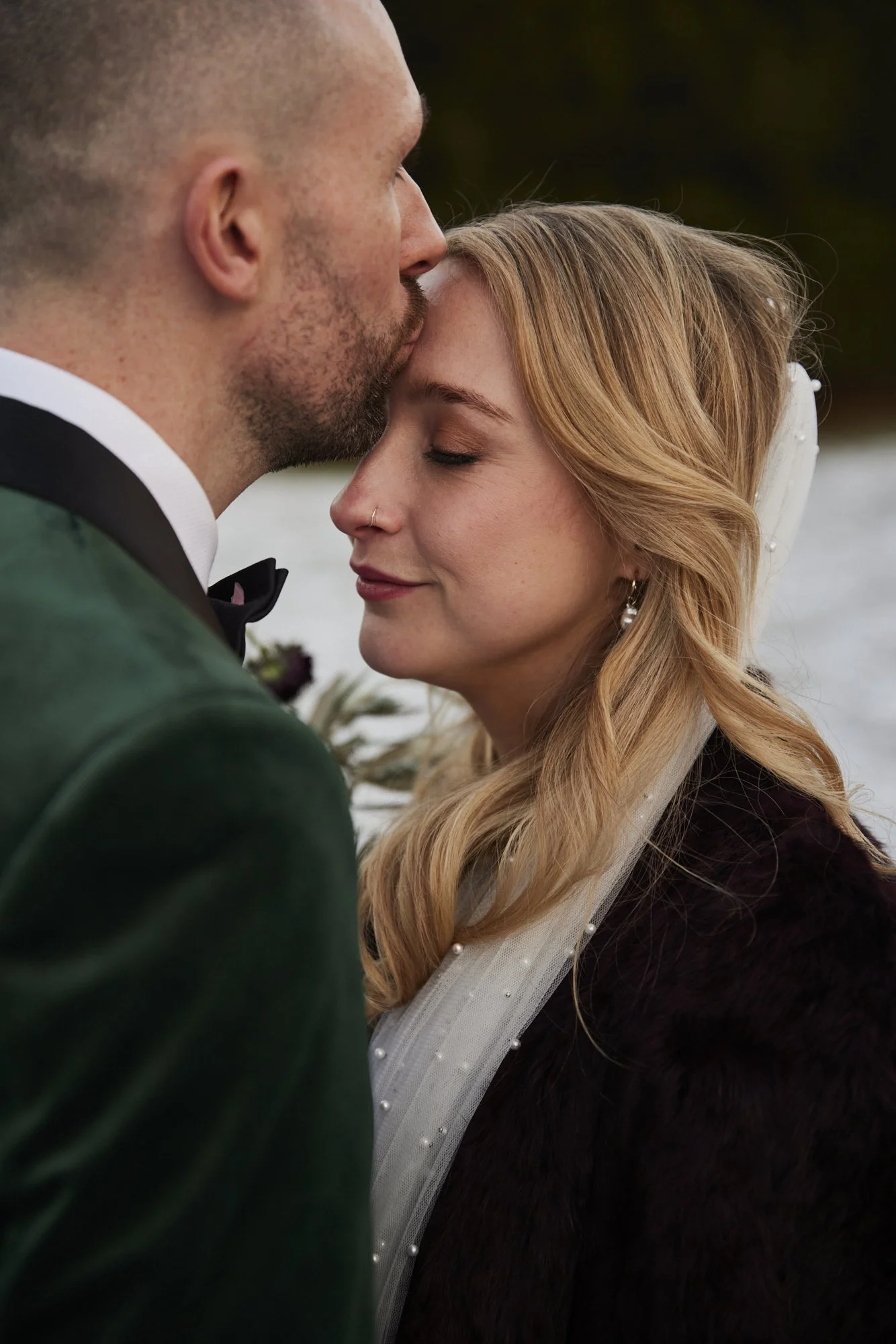 A man with a beard and a woman with blonde hair, both dressed in formal attire, sharing an intimate moment, with the man kissing the woman's forehead.