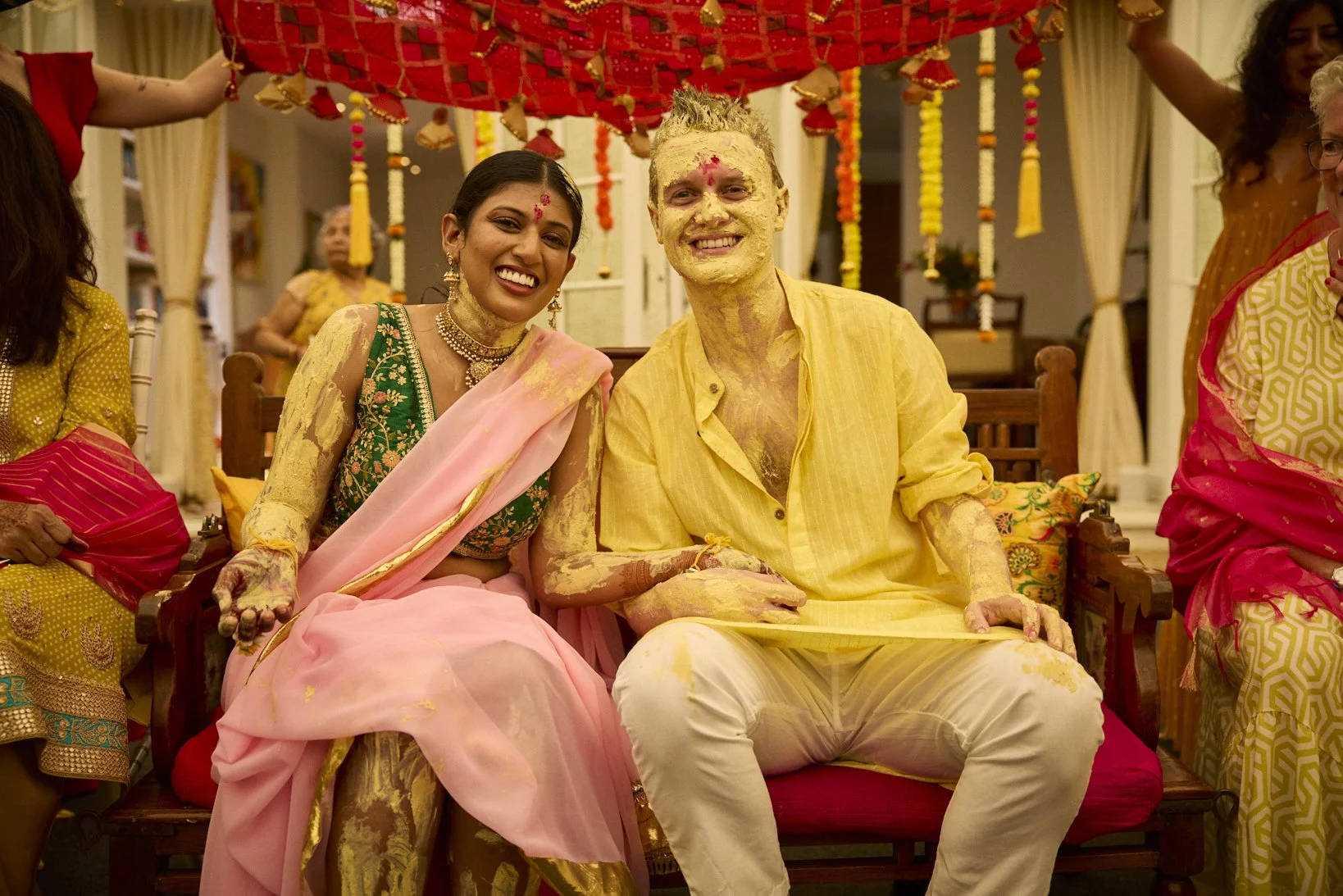 A couple sitting on a wedding bench, celebrating a festival with yellow powder on their faces and clothing, surrounded by other women in colorful traditional attire under a decorated canopy.