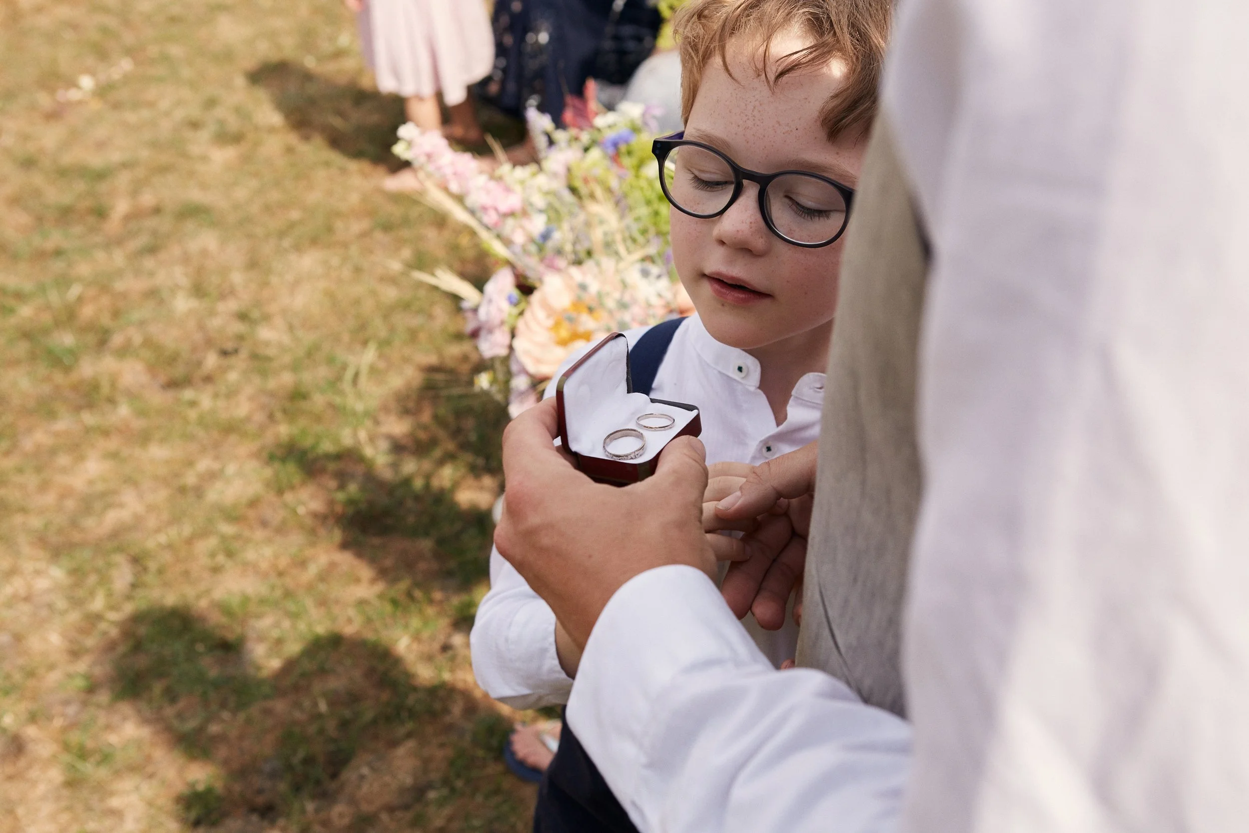 A young boy with glasses is receiving a ring from an adult, who is holding a ring box, at an outdoor wedding ceremony. There are flowers and other guests visible in the background.