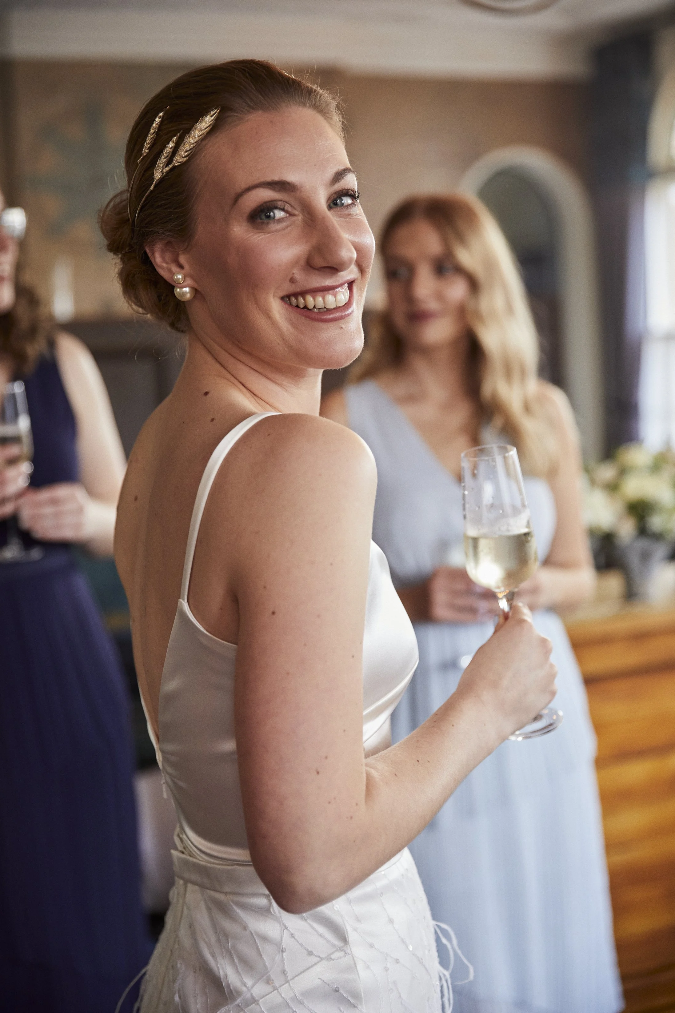 A woman in a white satin dress holding a glass of champagne, smiling at the camera during a celebration, with other women in the background.