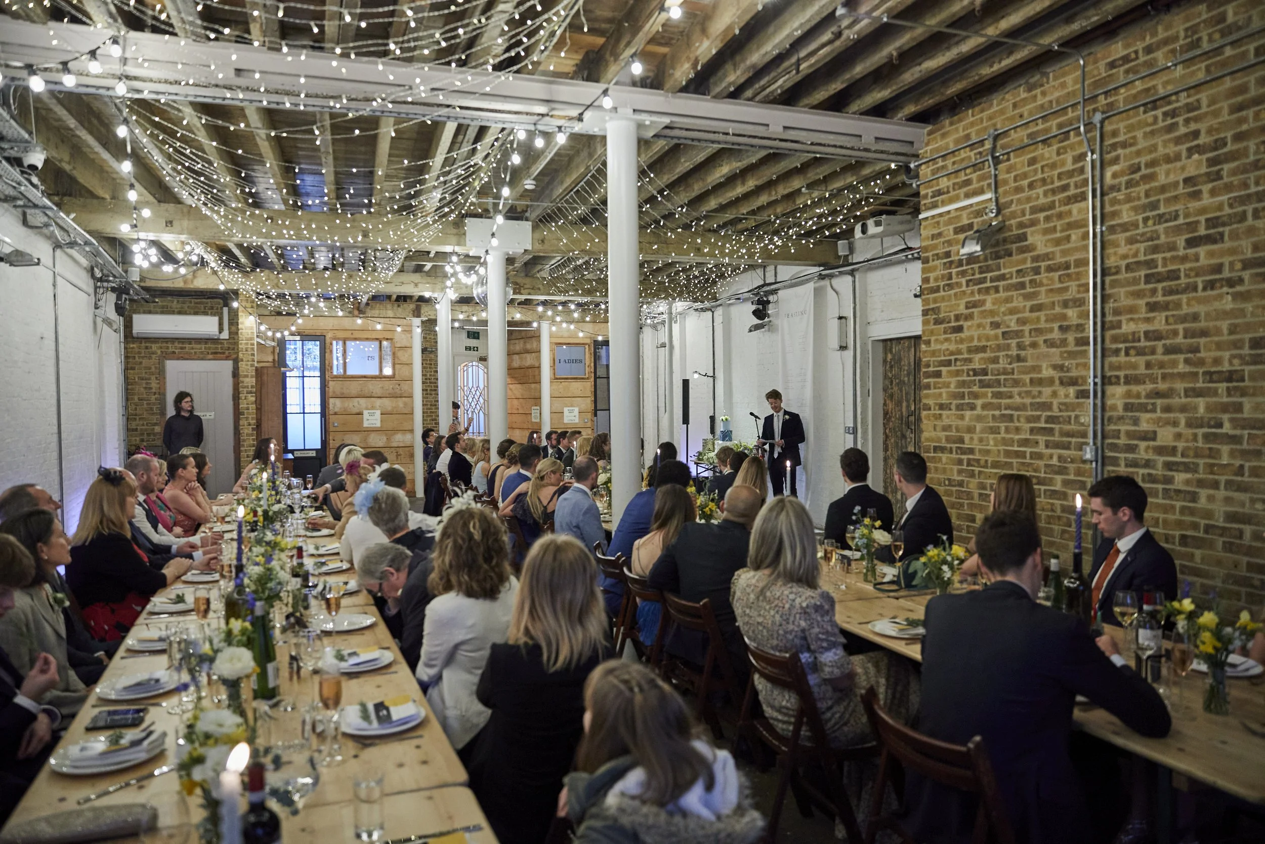 A large indoor wedding reception with long decorated tables, guests in formal attire, and a speaker on stage with a microphone, under string lights hanging from the ceiling in a rustic-style venue with exposed brick walls and wooden beams.