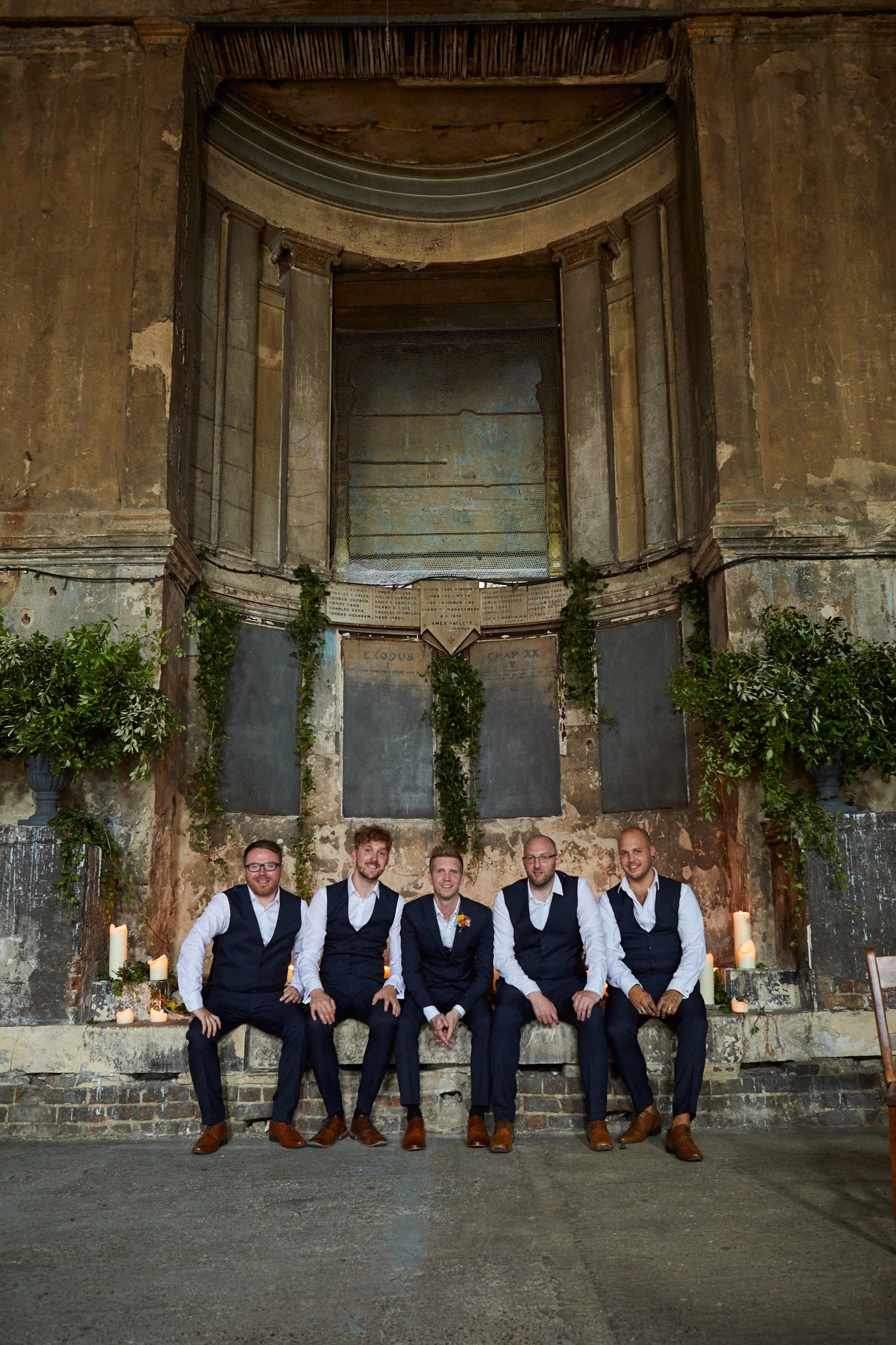 Five men in formal wedding attire sitting on a low ledge inside an old, abandoned church with aged walls, candles, and greenery.