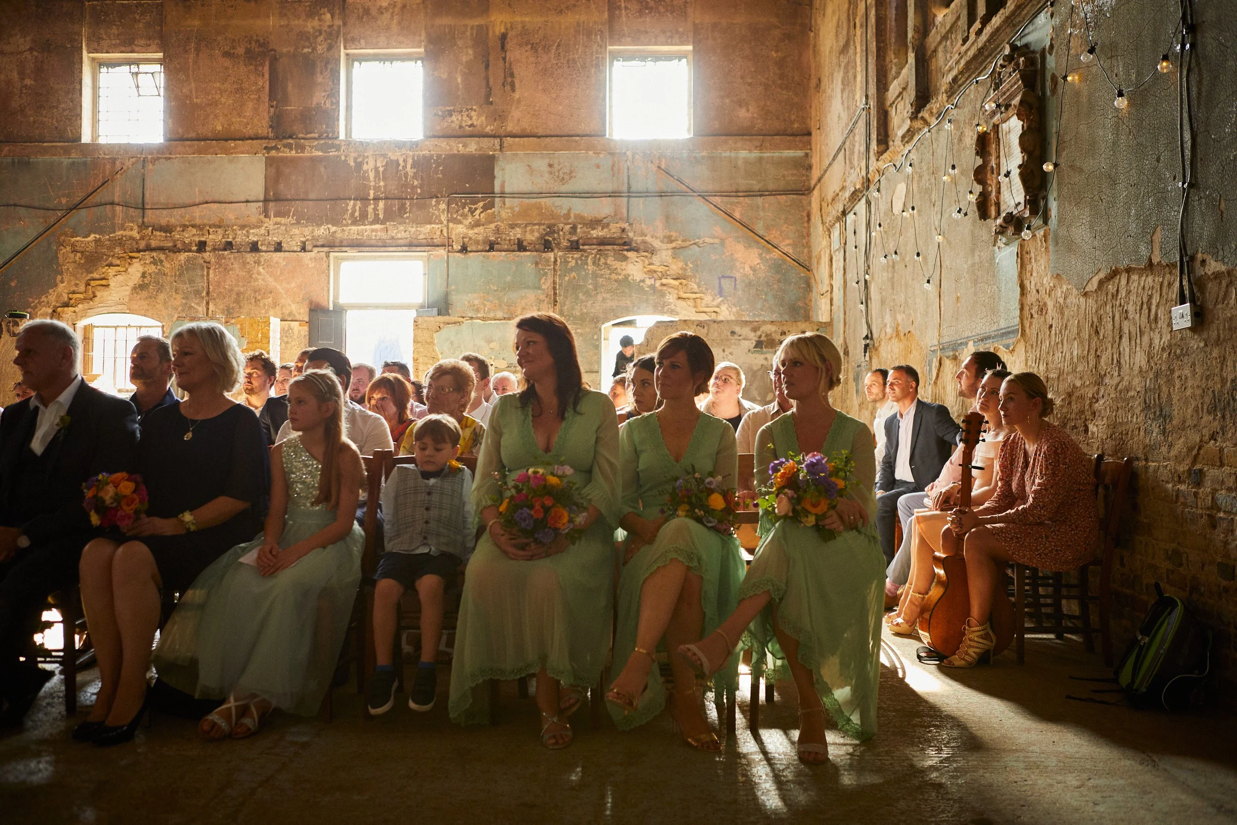 People attending a rustic wedding ceremony sit on wooden chairs inside a dilapidated building with large windows and string lights.